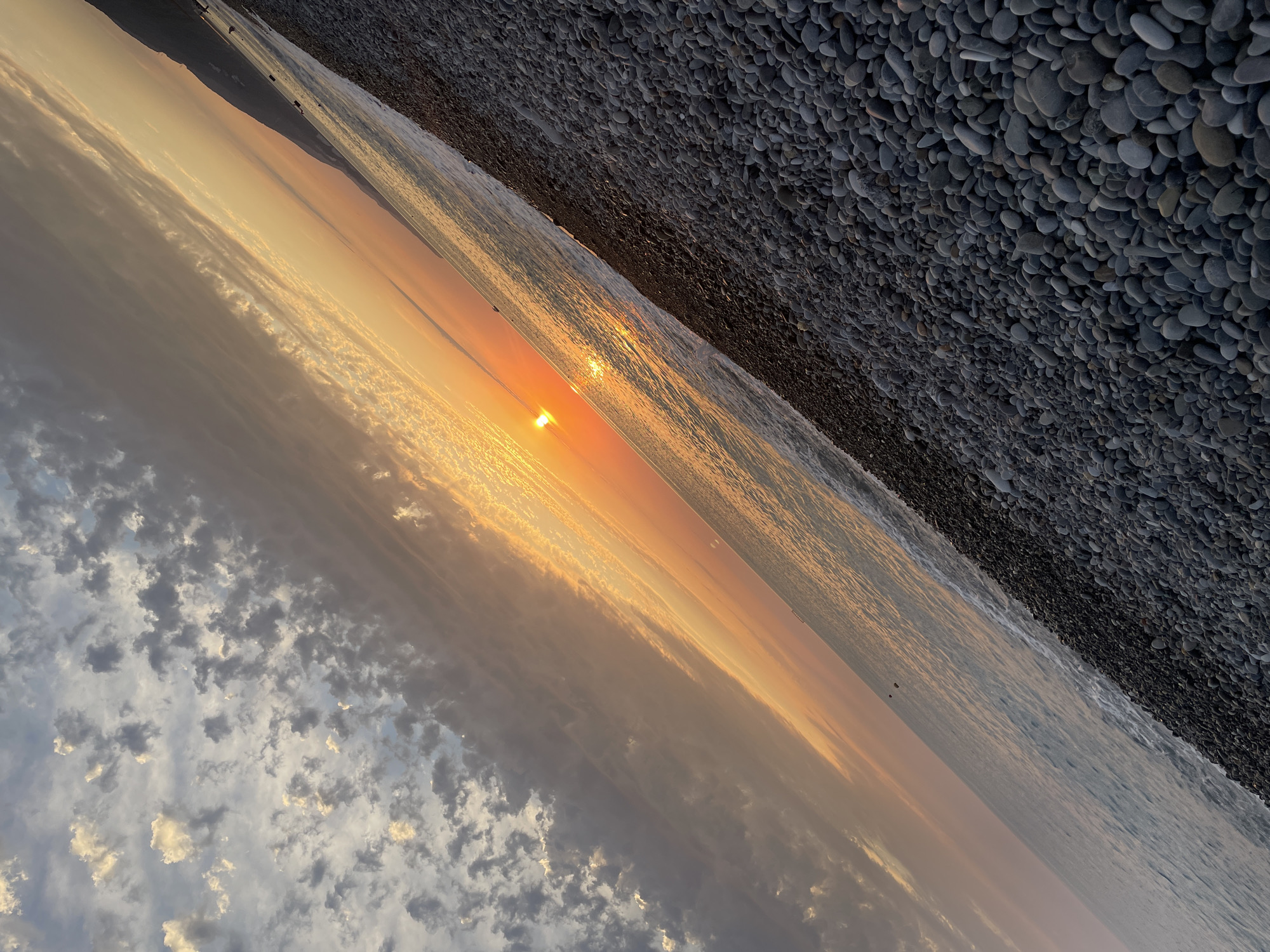 This is a photograph of a beach with a sunset. The image is taken from a low angle, with the camera tilted. The foreground is dominated by a densely packed field of grey and black pebbles that make up the beach. The pebbles are of various sizes and shapes, some smooth and rounded, others more angular. To the left of the pebbles is the sea, its surface reflecting the colors of the sunset. Gentle waves are breaking on the shore.
In the distance, a thin strip of land, likely a coastline or mountains, is visible against the bright sky. The sky is a canvas of warm hues, with the sun a brilliant orange orb, low on the horizon, casting a golden glow across the scene. Wispy clouds, tinged with orange and grey, stretch across the sky. The overall impression is one of a peaceful and serene sunset at the seaside.