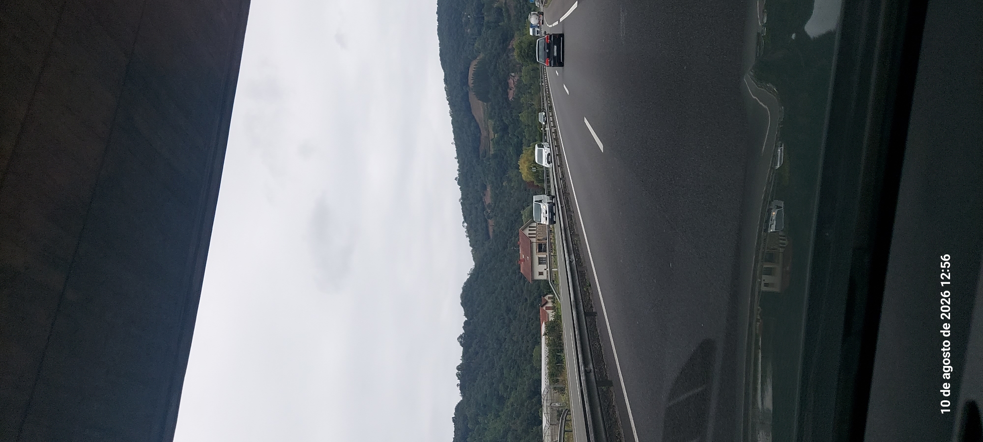 The image captures a highway scene with a cloudy sky over rolling green hills. In the foreground, the asphalt road is visible, with multiple lanes marked by white lines. Several vehicles are present on the highway, including a black Volkswagen Golf in the right lane, two white vans in the left lanes, and other cars further in the distance. On the left side of the highway, a metal guardrail separates the road from the adjacent landscape. Beyond the guardrail, there are some buildings, including a house with a red roof and a greenhouse structure. The background consists of densely wooded hills. At the bottom left of the image, a timestamp reads "10 de agosto de 2026 12:56". While the image is taken from a moving vehicle, there are no visible people. The scene appears to be an outdoor, daytime shot of a highway in a rural or semi-rural area. The location of Zumaia, Spain is not directly evident from the visual elements in the image itself, but is provided as context.