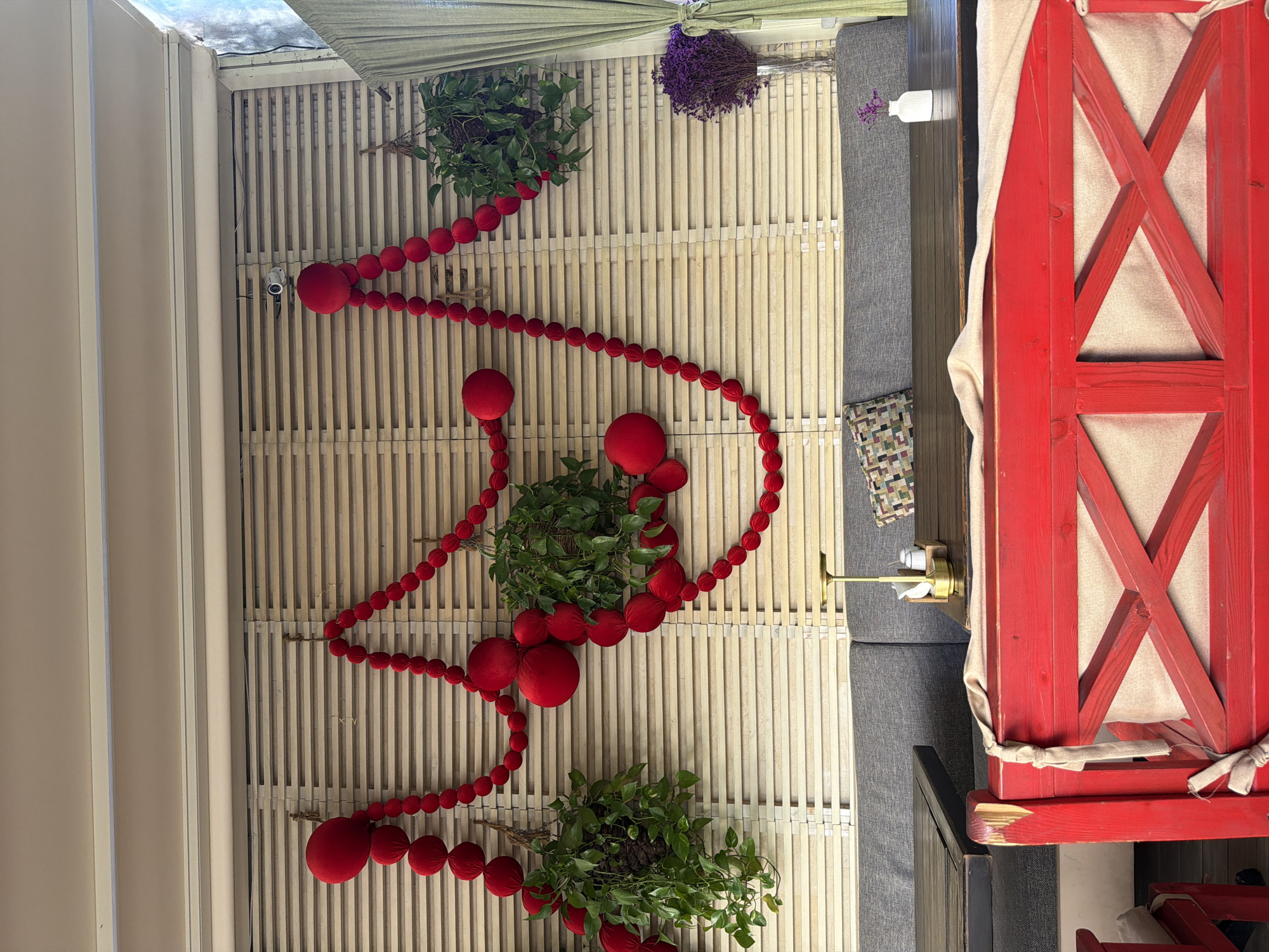 The image shows an outdoor seating area of a cafe or restaurant, likely in Khimki, Russia, given the context. The wall behind the seating is made of light-colored wooden slats. Two hanging potted plants with green leaves are suspended from the wall, partially obscured by red, spherical decorations strung together in a garland. The garland is draped across the wall in a decorative pattern.
A gray upholstered bench runs along the wall, with dark wooden tables in front of it. On one table, there's a small white vase with dried purple flowers and a salt and pepper shaker set on a brass stand. A patterned cushion is placed on the bench.
In the foreground, a bright red wooden bench with a rustic design is visible. It has a light-colored fabric cushion. The overall impression is of a cozy, decorated outdoor dining space. There are no people visible in the image.