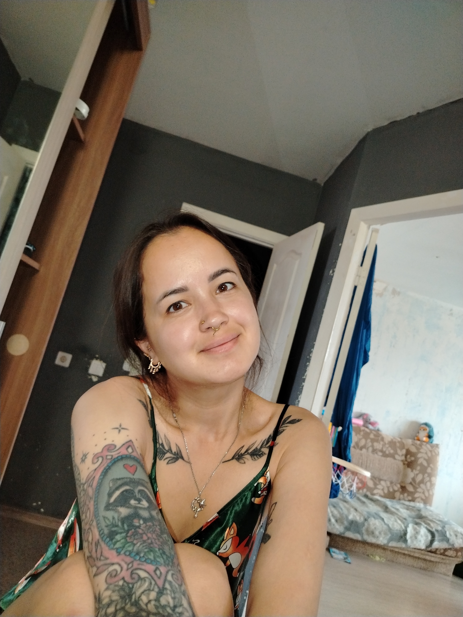 The image is a medium shot of a young woman with dark hair and brown eyes, smiling at the camera. She is wearing a green slip dress with a fox pattern and a silver necklace with a heart-shaped pendant. She has several tattoos on her arms and chest, including a raccoon in a frame on her left arm. She is also wearing a septum piercing and gold hoop earrings. The woman is indoors, in what appears to be a bedroom. The walls are dark gray, and there is a wooden wardrobe to the left of the frame. To the right, there is an open doorway leading to another room with a bed and a stuffed animal. The lighting is natural and soft, coming from a window or light source not visible in the frame. The image quality is moderate, with some noise and blur visible. The context of Noviy, Russia, is not evident from the image itself, as there are no specific geographical markers or cultural elements that would indicate this location.