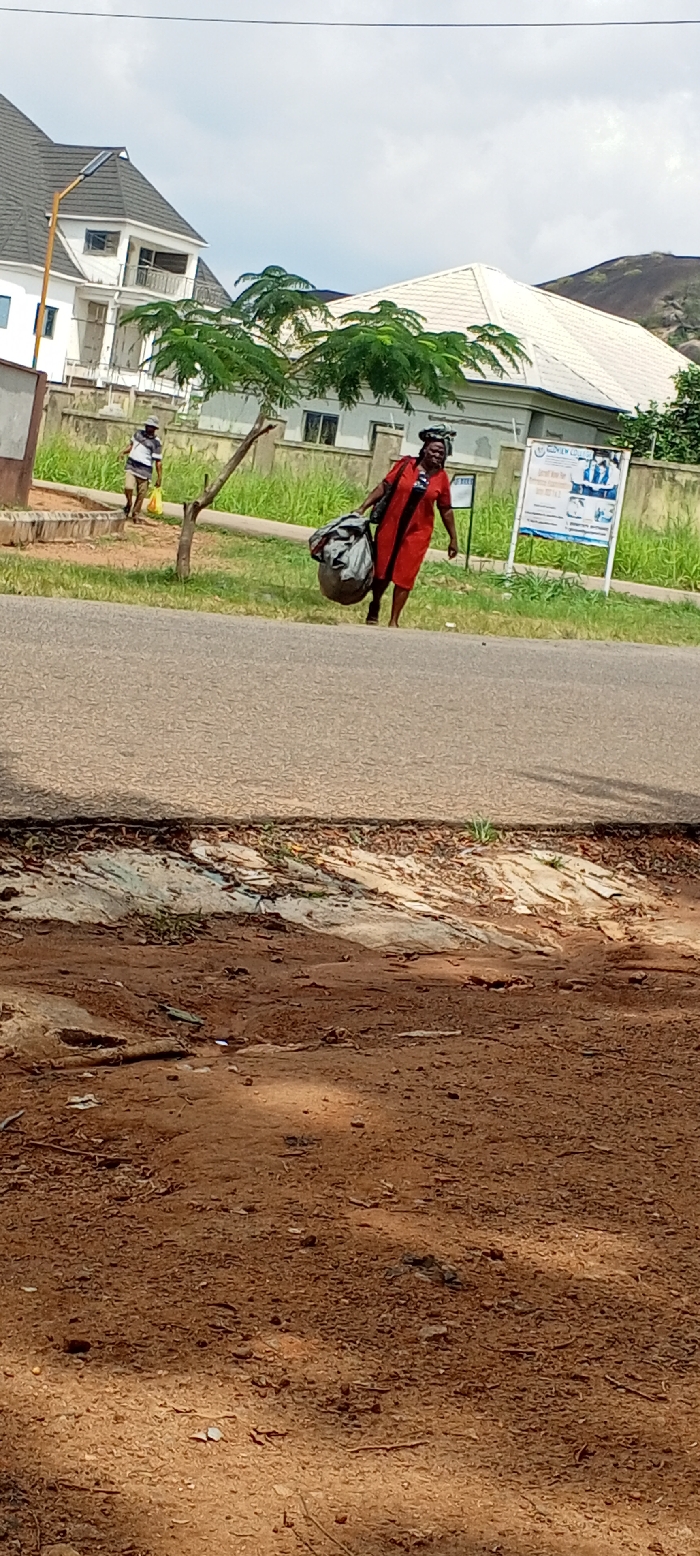 This image depicts a daytime outdoor scene along a paved road in Akure, Nigeria.
In the mid-ground, a dark-skinned adult female is walking from right to left, parallel to the road. She wears a red dress or tunic over a patterned garment and has an object or headwrap on her head. She carries a large, dark, bulging bag or bundle over her left shoulder. Further in the background on the left, an adult male walks from left to right, wearing a striped shirt, dark pants, and a light-colored hat, carrying a yellow plastic bag in his right hand.
The paved road occupies the lower-middle section of the frame. Beyond the road is a dirt verge leading to a grassy area. A slender tree with green leaves stands between the two individuals. To the right, a white signboard with blue text and graphics, displaying "MIDAS ACADEMY" and "ADMISSION FORMS," is visible within the grassy area.
In the background, behind the man, is a large, multi-story white building with a dark-tiled, multi-pitched roof and balconies. A yellow pole with a solar panel is attached to its side. Further right, behind the signboard, another building with a light-colored, pitched roof is partially visible. Dark, rocky hills are present in the far background on the right, under a partly cloudy sky. The immediate foreground consists of reddish-brown dirt with scattered debris.