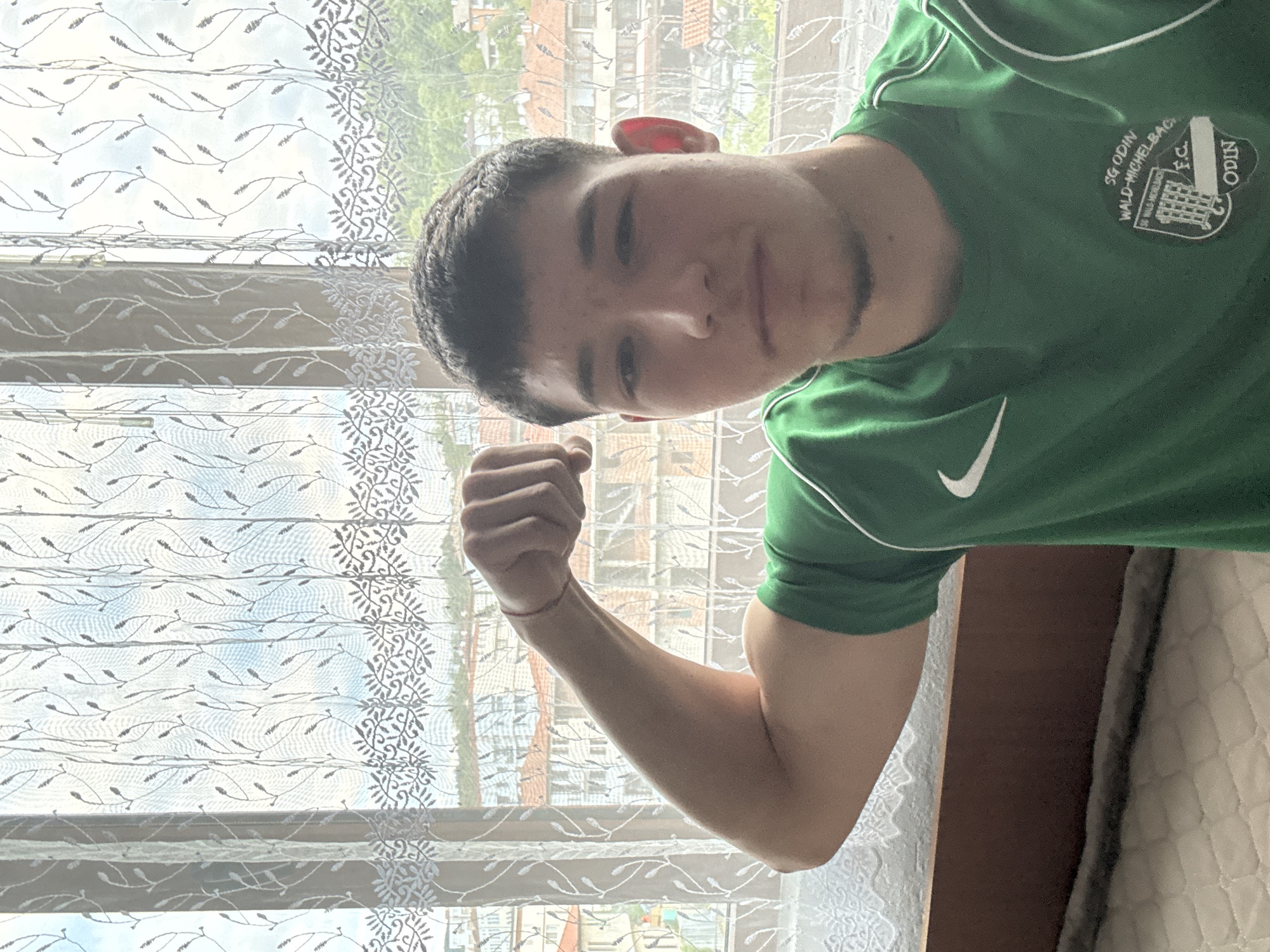 A young man is posing for a selfie indoors, wearing a green t-shirt with white trim and a logo on the left breast. He is flexing his right arm, displaying the bicep. A red bracelet is visible on his wrist. The background shows a window covered with a sheer, patterned curtain. Outside the window, buildings are visible. Based on the shirt logo, the photo may have been taken in or associated with Germany. No evidence suggests the location is Zlatograd, Bulgaria.