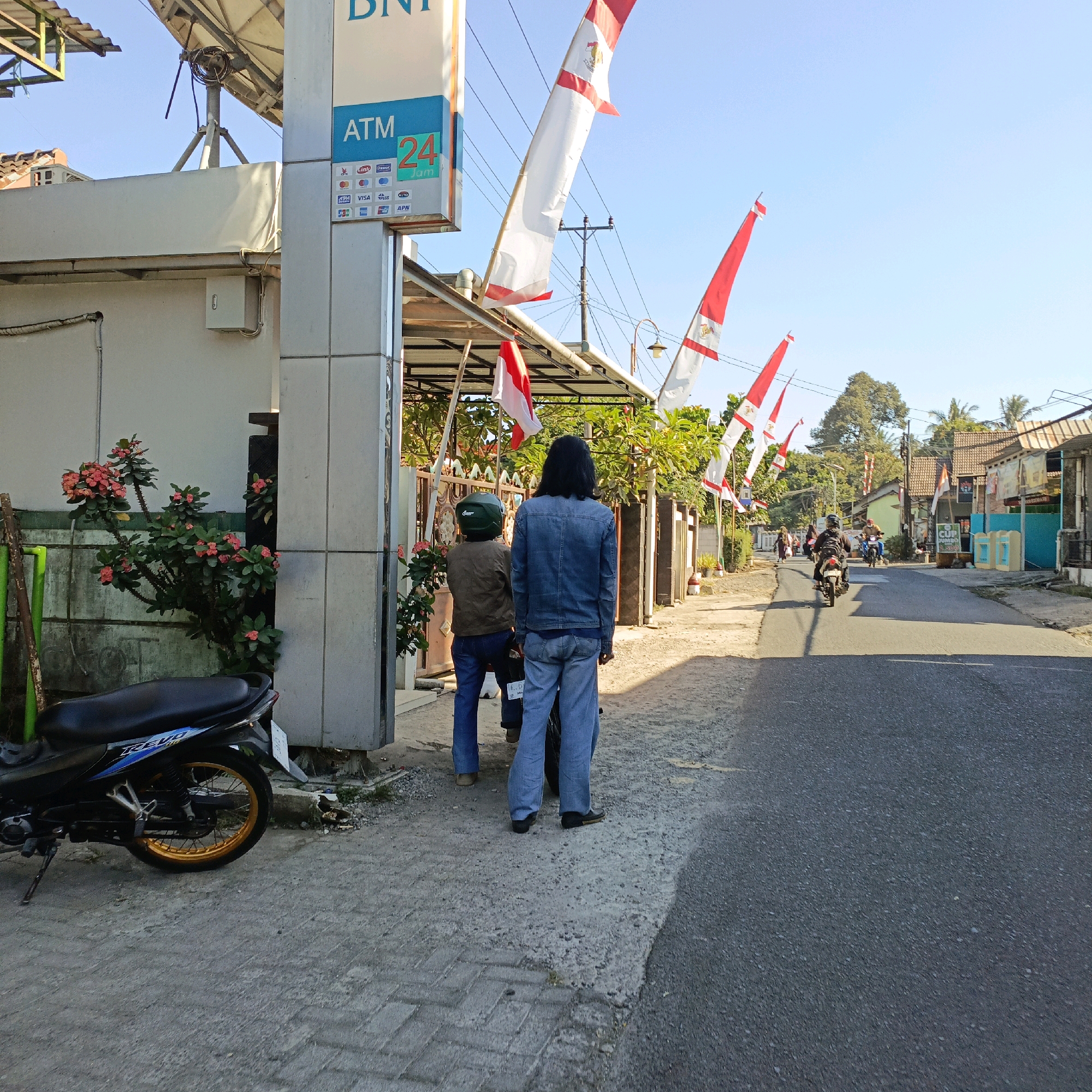 The image depicts a street scene in Jombor, Indonesia, bathed in bright sunlight under a clear blue sky. In the foreground, a motorcycle is parked on a paved sidewalk next to an ATM. Two individuals, seen from the back, stand near the ATM. One person wears a denim jacket and blue jeans, while the other is dressed in a brown jacket and blue jeans, with a green helmet. Several Indonesian flags, red and white, are displayed, some hanging from poles and others from a canopy structure. The ATM sign indicates it is for BNI bank and operates 24 hours. The street itself is paved and shows signs of light traffic, with a few motorcycles visible further down the road. Buildings line both sides of the street, featuring a mix of architectural styles. Lush green trees and foliage are present, particularly to the right of the ATM and along the street. The overall impression is of a typical day in a local Indonesian community.