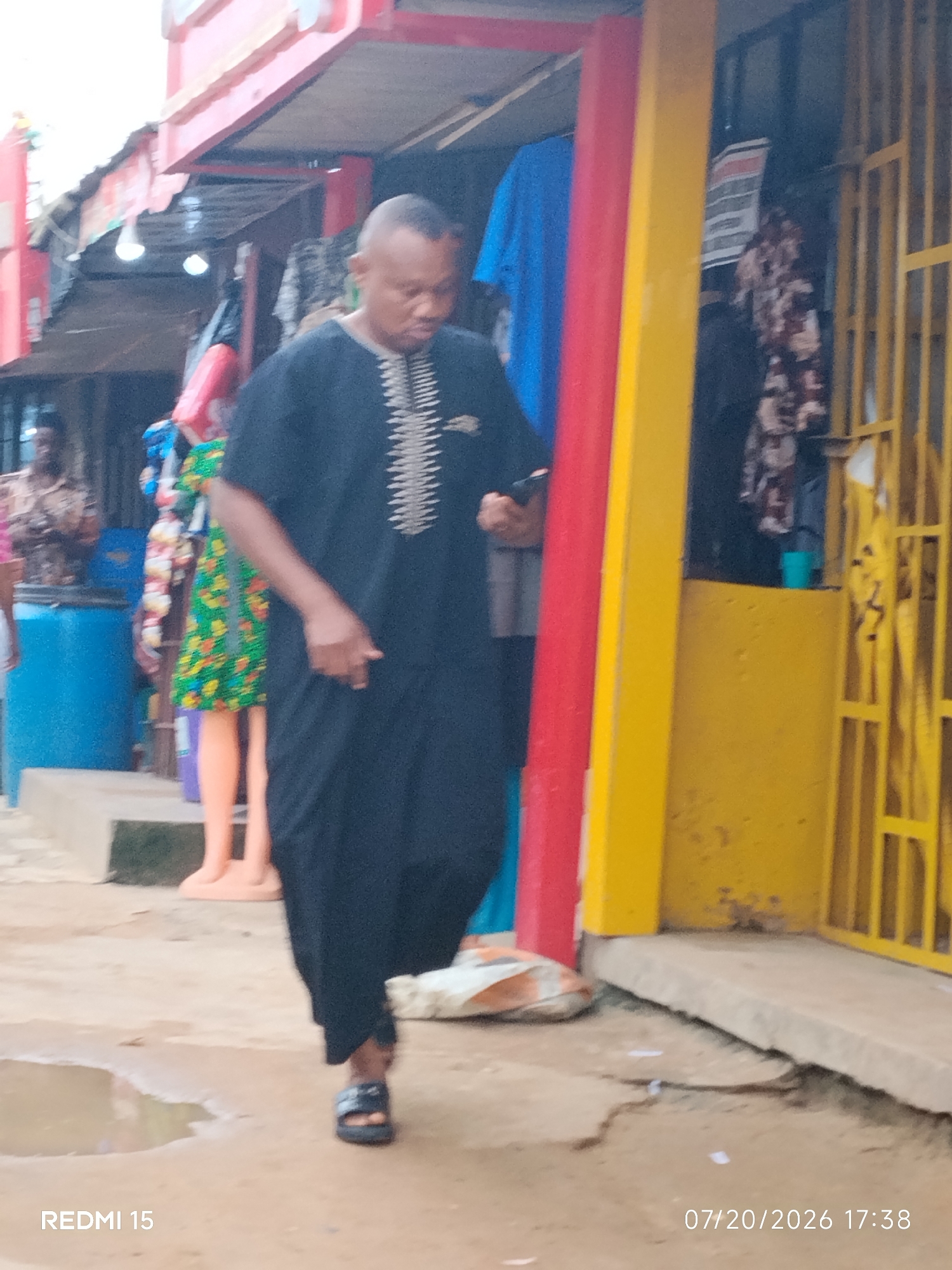 The image, dated 07/20/2026 at 17:38, depicts a street scene in Umuode, Nigeria. A man in a black traditional outfit with white embroidery is walking towards the right side of the frame. He is wearing dark sandals. In the background, there are shops or market stalls with colorful displays of goods. To the left, a blue barrel and a mannequin wearing a green, floral-patterned dress are visible. A red and yellow pillar stands prominently in the center of the image, separating different sections of the market. The ground is a dirt path with some puddles. The overall impression is of a bustling market environment.