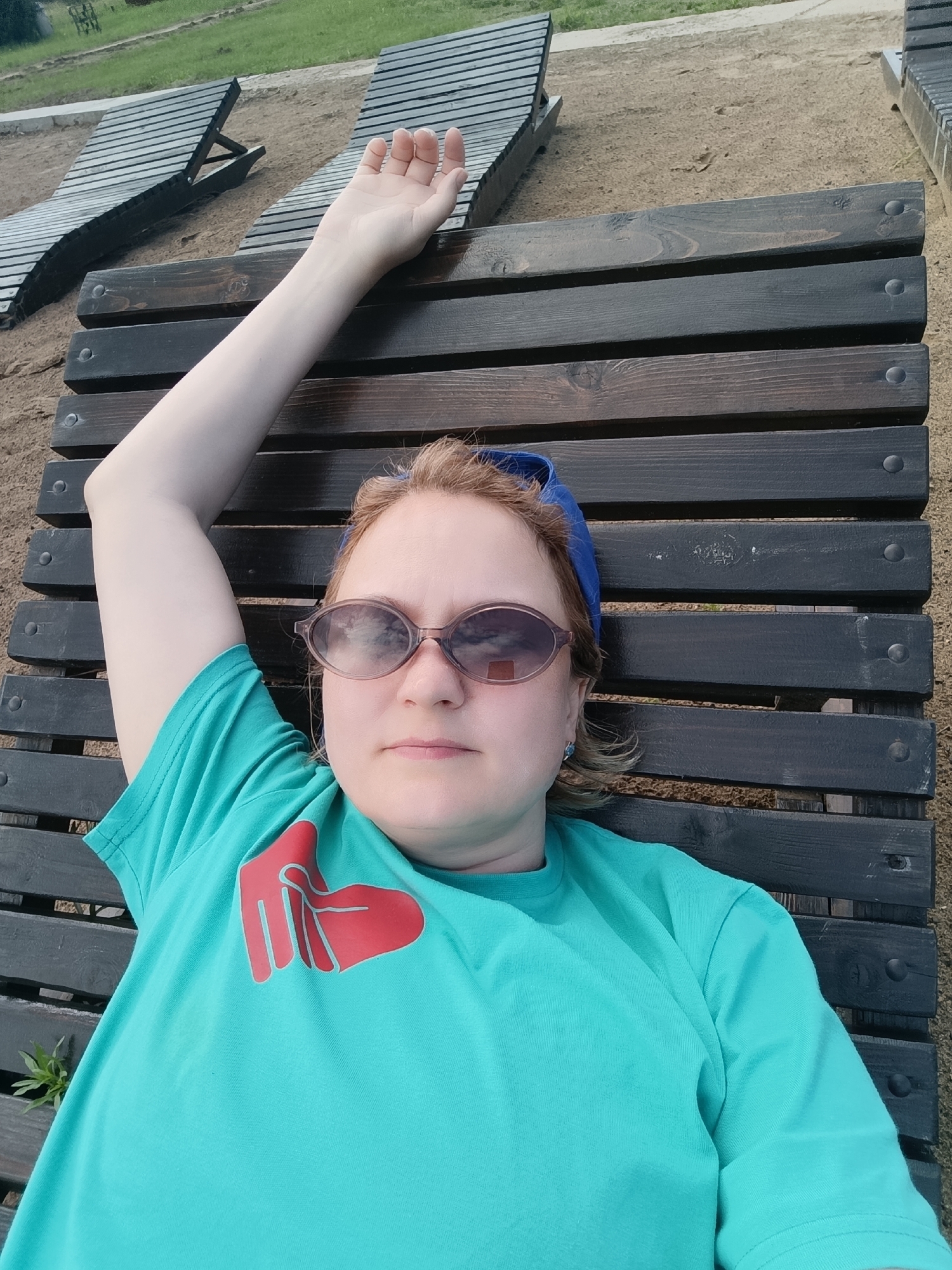 A young woman with light brown hair and sunglasses is lying on a wooden lounger. She is wearing a teal t-shirt with a red heart-shaped graphic on it and a blue headscarf. Her left arm is raised above her head, and her right arm is bent, resting on her chest. The lounger is made of dark wood planks and is set on a sandy area. In the background, there are more wooden loungers and a grassy area. The location is Prikamskii, Russia.