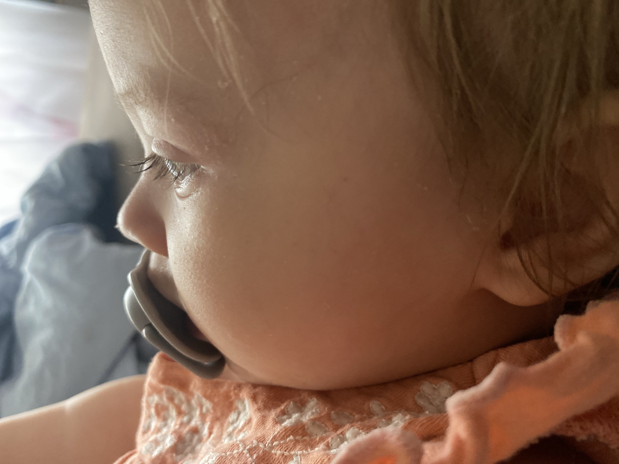The image is a close-up, profile shot of a baby or young toddler, captured from the right side, looking towards the left. The subject's face dominates the frame, showcasing their delicate features.
The child has fair skin and fine, light-colored hair, possibly blond or light brown, visible around the forehead and temples. Their eyelashes are notably long and appear slightly clumpy or wet, enhancing the focus on the eye area. A small nose and chubby cheeks are visible. Small, light-colored flakes or particles are noticeable on the child's skin, particularly on the forehead and near the eye, and also in their hair.
A gray pacifier is inserted into the child's mouth, with the handle and shield visible protruding from the mouth. The child's earlobe is partially visible behind the pacifier.
The child is wearing an orange or peach-colored garment, possibly a onesie or dress, which features a white, textured or embroidered floral/patterned design around the neckline and shoulder area. A bare arm is partially visible below the shoulder.
The background is softly blurred, indicating a shallow depth of field. To the upper left, a bright, diffused light source or a light-colored object (like a sheet or wall) creates a soft, luminous effect. Below the child's head and behind their back, a blurry dark blue or gray fabric is visible, possibly a blanket or another item of clothing.
The lighting appears soft and natural, emphasizing the contours of the child's face. The overall impression is one of peaceful observation or rest, capturing a tender moment. There is no visible text in the image.