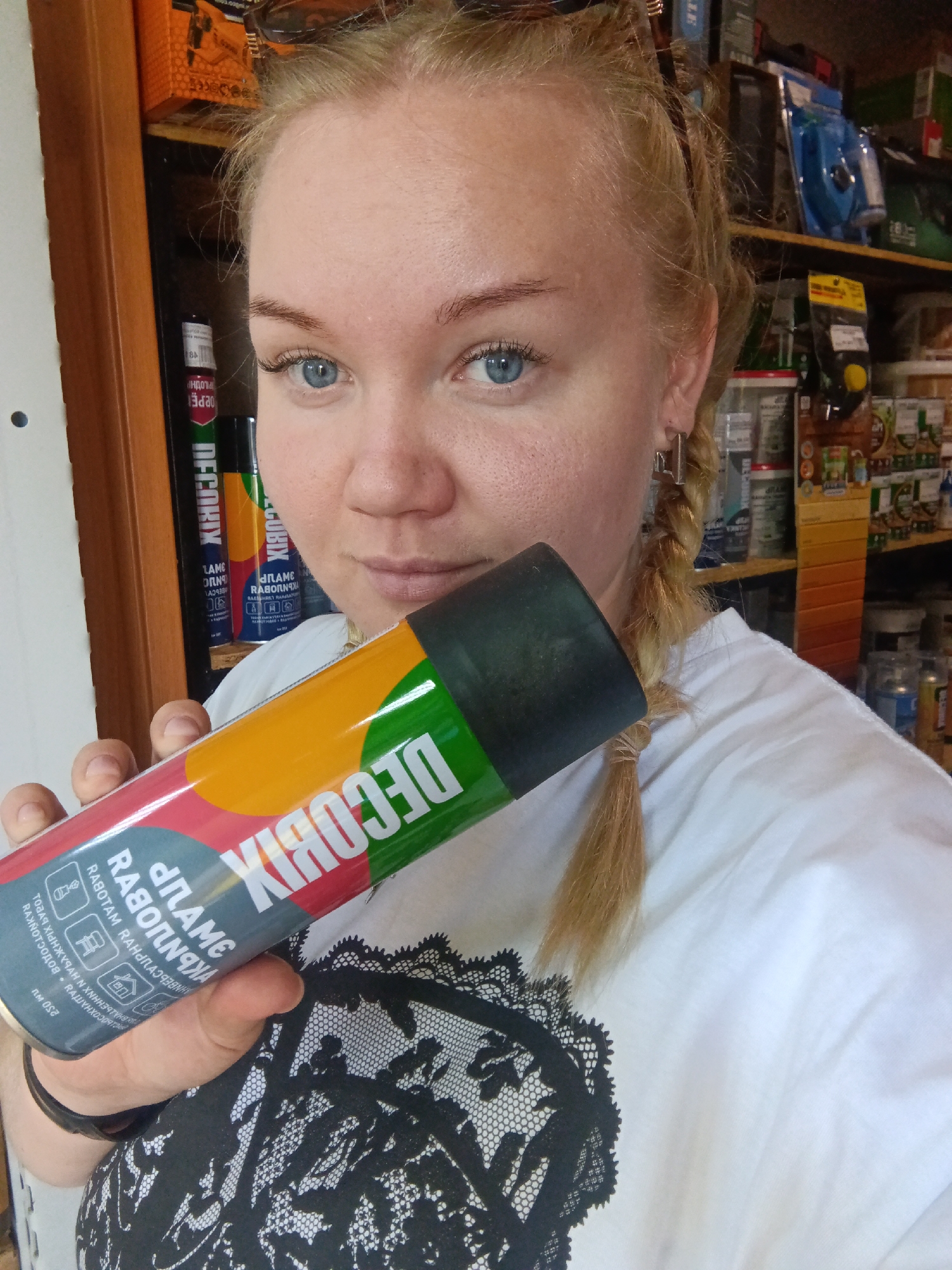 The image shows a woman holding a can of DECOBIX spray paint. She is standing indoors in what appears to be a hardware or general store, judging by the shelves stocked with various items in the background. The woman has blonde hair styled in braids and is wearing a white t-shirt with black lace detailing. The can of DECOBIX spray paint features a multi-colored design with red, orange, yellow, and green sections. The label includes text in Russian. The location is likely in the city of Фоки, Russia, based on the text on the can.