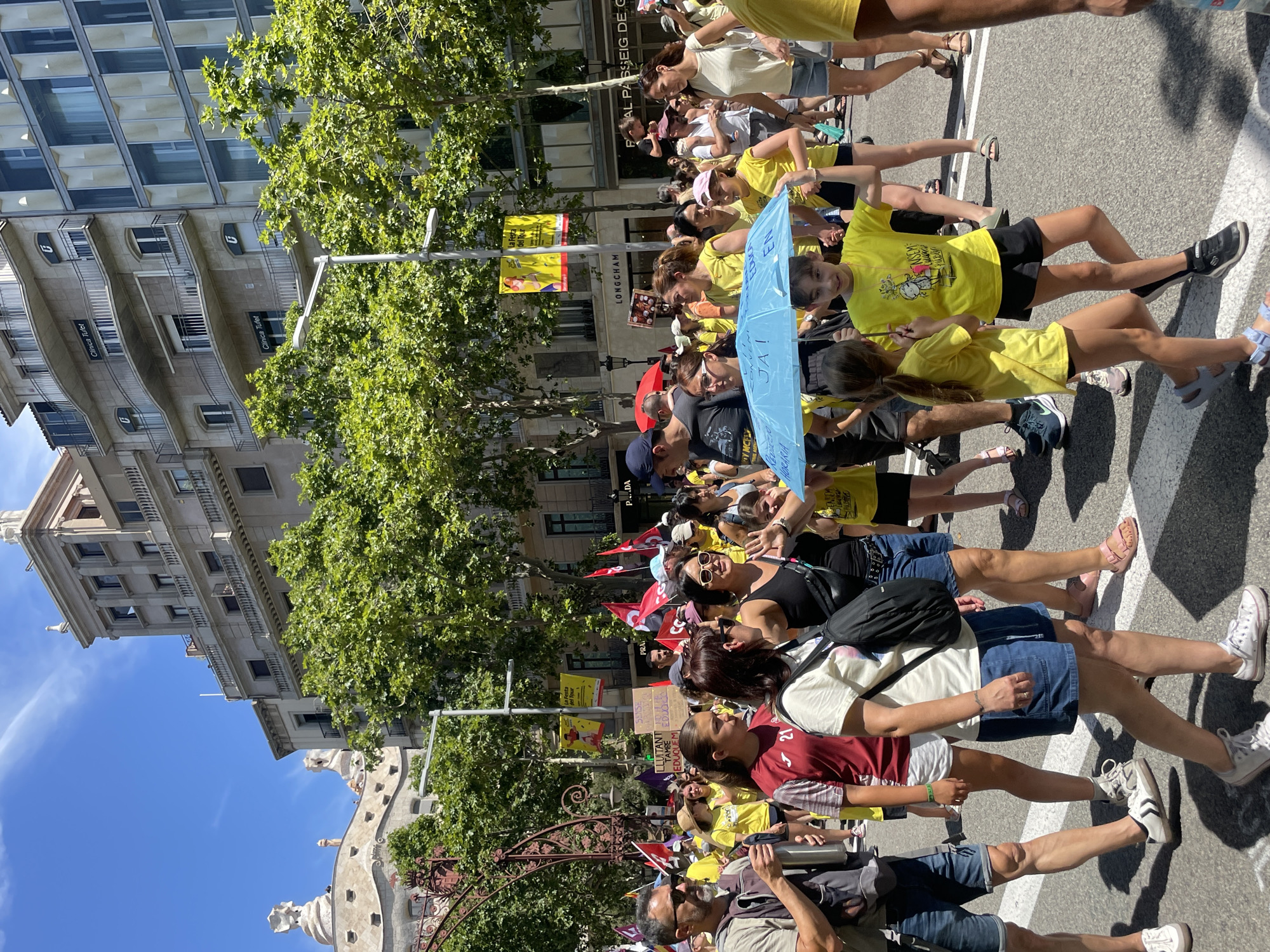 The image shows a street scene in Barcelona, Spain, likely during a protest or parade, given the presence of flags, signs, and a large gathering of people. The people, predominantly dressed in yellow and other casual attire, are walking along a street lined with trees and multi-story buildings. Some individuals are holding flags, including red flags with symbols, and banners with text. One prominent banner in the background reads "La Fira del Tur" in yellow lettering on a red background. In the foreground, a young girl holds a bright blue umbrella over herself and another child. The architecture of the buildings suggests an urban setting, with balconies and ornate facades. The sky is clear and blue with some wispy clouds. The overall atmosphere appears to be lively and engaged, with people of various ages participating in the event.