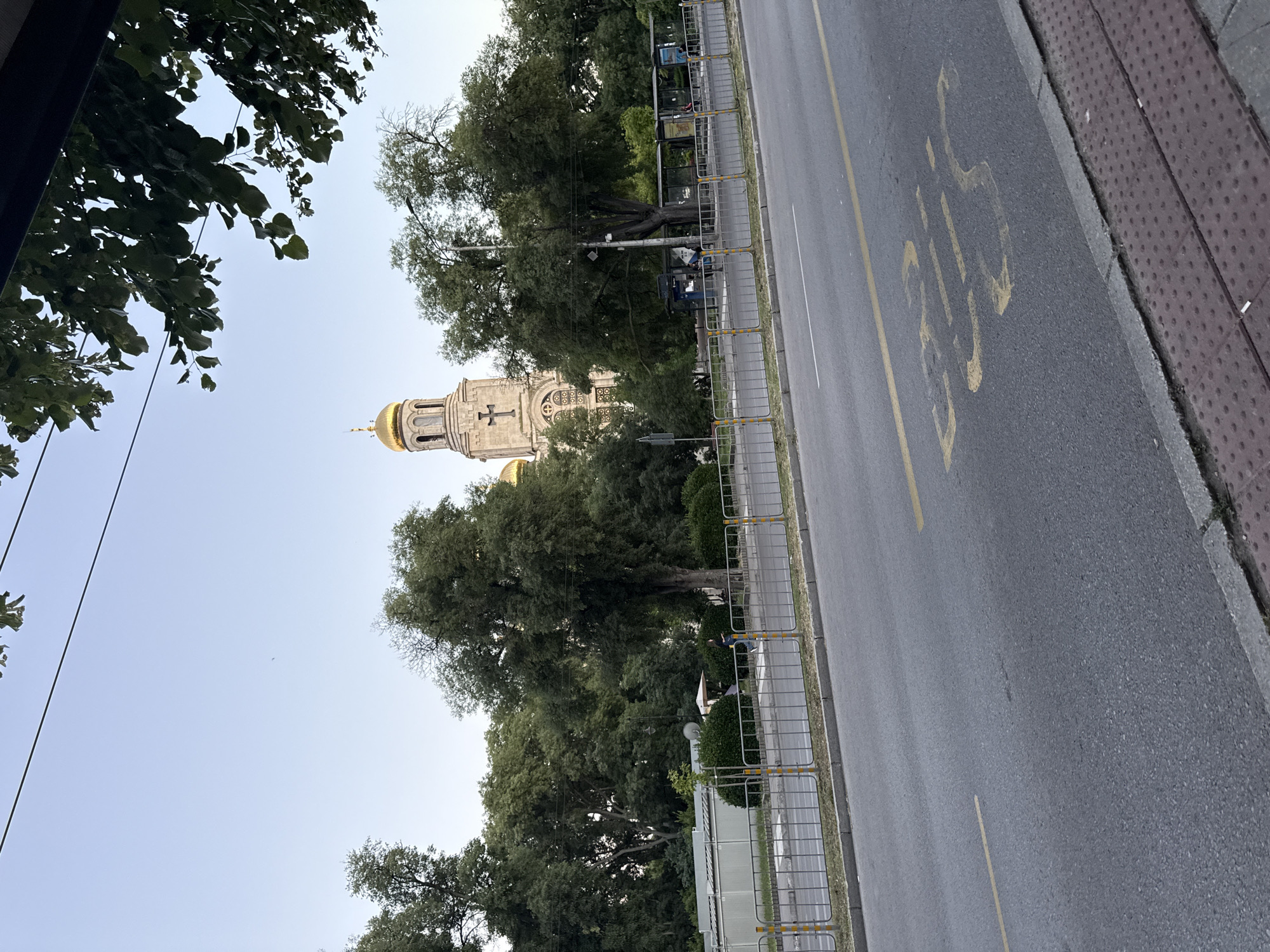 The image captures a street scene in an urban environment, likely during the day given the lighting. In the background, a large building with a golden dome and a cross is visible, suggesting it is a religious structure, possibly a cathedral. This building is surrounded by numerous mature trees, creating a green canopy. The foreground shows a paved road with yellow lane markings, including a designated bus stop area marked with yellow paint. A sidewalk with textured paving is visible to the right of the road. Beyond the road and a metal fence, there are bus shelters and people are discernible near them, waiting or passing by. The sky is clear and light blue. The overall scene depicts a public space with elements of architecture, nature, and urban infrastructure.