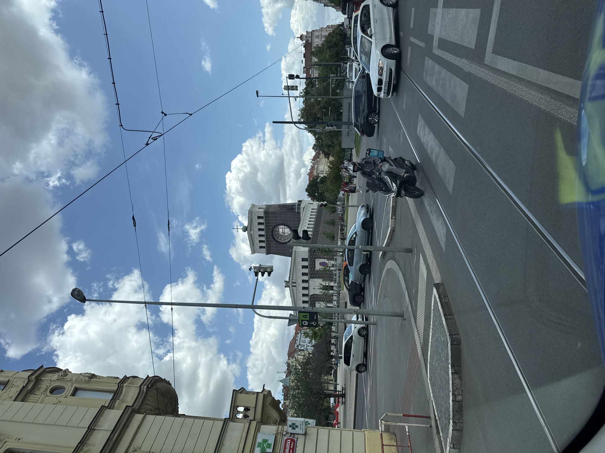 The image captures a street scene in Prague, Czechia. A streetcar is visible in the foreground, partially obscured by its window. The street is paved with asphalt and features tram tracks. Several cars, including a white hatchback and a dark sedan, are on the road, some stopped at a traffic light. A scooter rider, wearing a helmet and a blue delivery bag, is navigating through the intersection.
In the background, a large, ornate building with a clock tower stands prominently. The architecture suggests a historical or significant public building. Other buildings with traditional European design are also visible. The sky is partly cloudy with white cumulus clouds against a blue backdrop. The environment appears to be a busy urban intersection with pedestrian crossings marked on the road. A street lamp and traffic signals are also present. A pharmacy sign is visible on the left side of the image, indicating a commercial area.