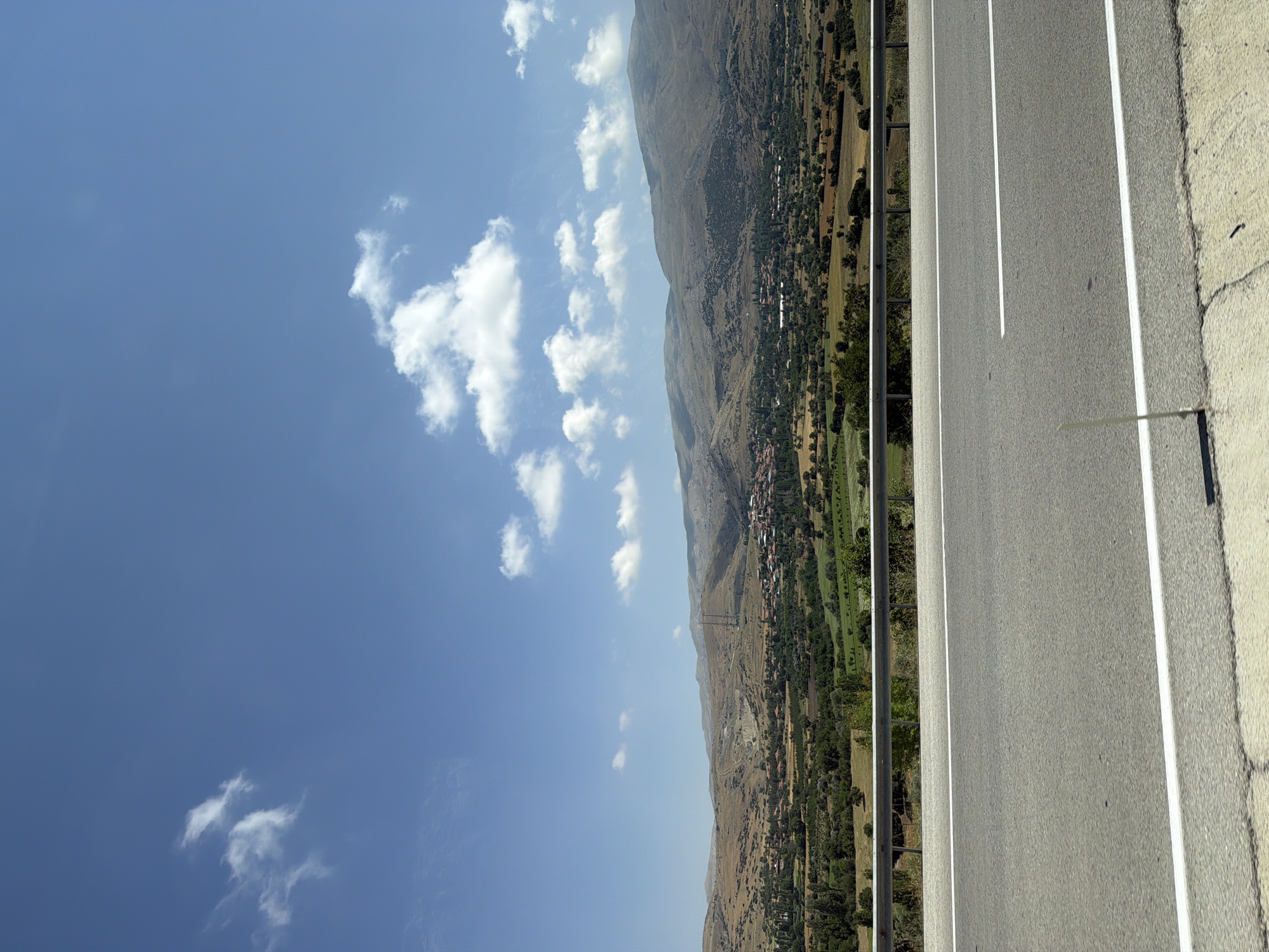 The image depicts a wide shot of a rural landscape under a bright blue sky with scattered white clouds. In the foreground, a paved road with white lane markings stretches horizontally across the frame. A metal guardrail separates the road from the embankment. Beyond the road, the landscape unfolds into rolling hills covered with dry, scrubby vegetation and patches of green fields. A small village, characterized by numerous red-tiled roofs, is nestled in the valley below, surrounded by trees. In the background, larger, more rugged mountains form a backdrop against the sky. The scene appears to be taken from an elevated vantage point, likely from a vehicle traveling on the road. The environment suggests a warm, sunny day in a semi-arid region. There are no people visible in the image. The location is identified as being in the vicinity of Çavdır, Turkey.
