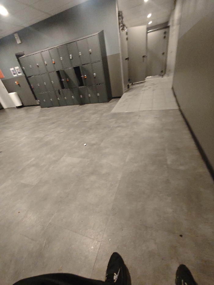 A gray-toned locker room or changing area. Lockers line the left wall. A door with a small window is visible in the background, hinting at a bathroom or shower area. The floor is gray tiled. In the foreground, the lower part of the view shows the person's legs and shoes. The lighting is muted and even, highlighting the utilitarian nature of the space. The room is empty.