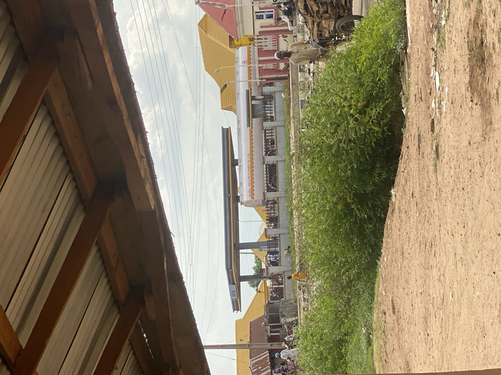 The image captures a street scene in Bali, Nigeria, under an overcast sky. In the foreground, a dirt path leads towards a dense thicket of green bushes. Beyond the bushes, a gas station with a prominent white tanker truck bearing the inscription "BALA MOHAMMED OIL LTD." is visible. The gas station canopy is labeled "MRS." To the left of the gas station, buildings with corrugated metal and yellow tiled roofs are present, and several people are gathered near them. In the mid-ground, a man in a tan tunic pushes a cart laden with firewood. The overall environment suggests a developing urban area with a mix of commercial and residential structures, and evidence of daily life and commerce.