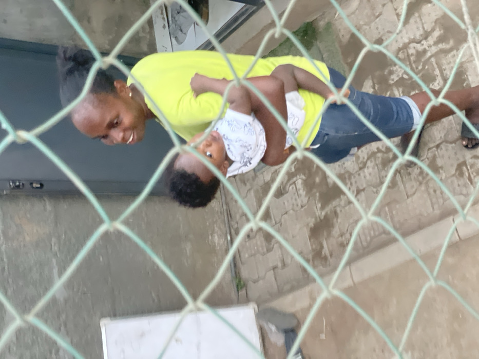 A woman in a neon yellow shirt and denim capri pants holds a baby wearing a patterned white and blue shirt. The woman has dark hair pulled back and is looking down at the baby. Both are dark-skinned. The image is taken through a green wire fence, slightly obscuring the subjects. The background is a paved area and part of a building with a door.