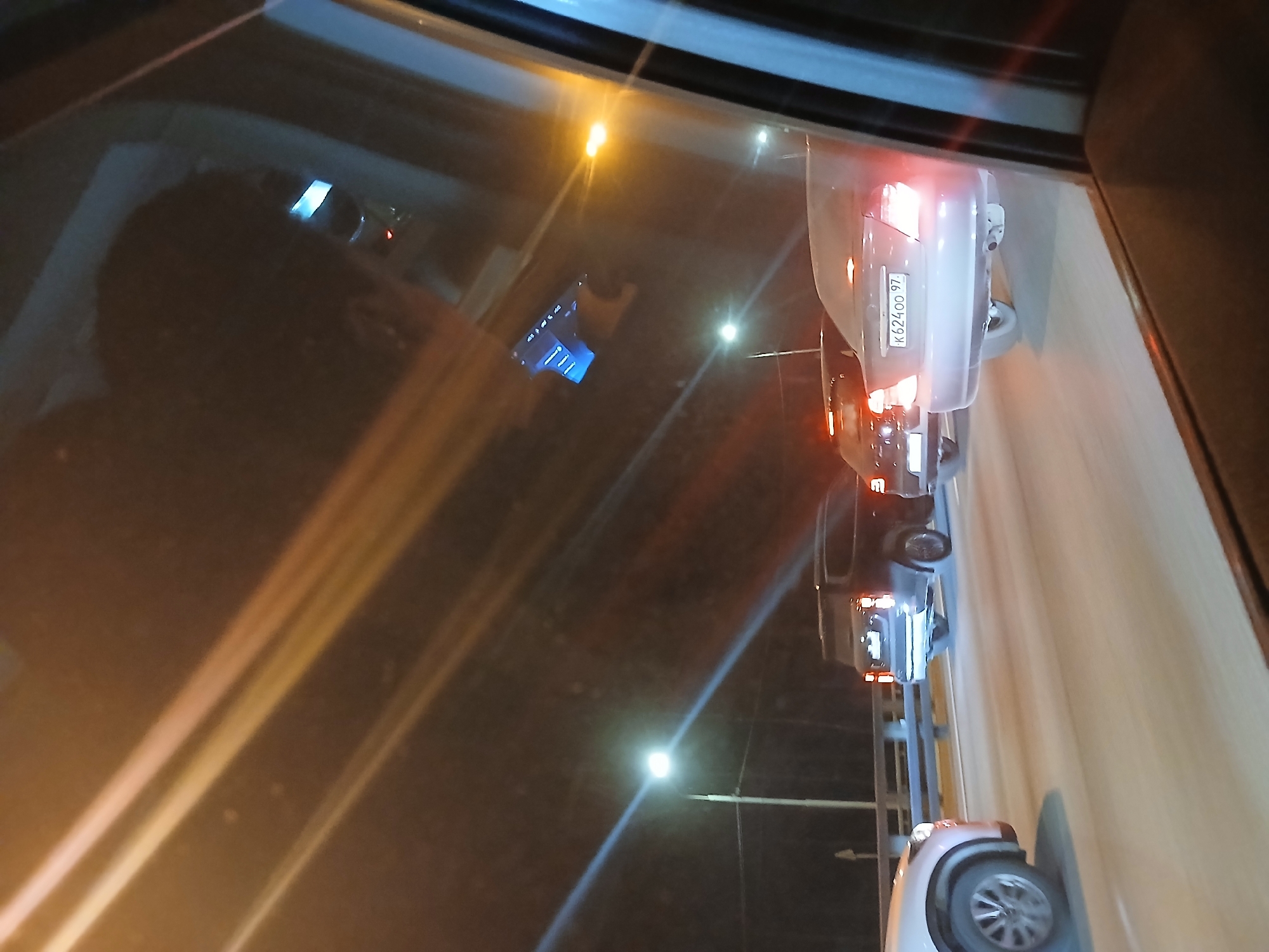 The image depicts a nighttime view from the interior of a vehicle, looking out a side window at traffic on a multi-lane roadway in the city of Unknown, Unknown. Several vehicles are visible, appearing to be in motion or stationary in traffic. In the mid-ground, a light-colored sedan displays a rear license plate reading "K 624 OO 97," with its taillights and brake lights illuminated. Directly behind it, a dark-colored SUV also has illuminated taillights. A third light-colored vehicle, possibly a sedan or SUV, is partially visible in the far left foreground. The road surface appears reflective, possibly damp. A metal barrier or guardrail runs along the left side of the road, suggesting an elevated road section or bridge. Bright streetlights on tall poles illuminate the scene from the left and right sides. Reflections of bright light streaks and a faint blue rectangular object are visible on the vehicle's window through which the image was captured. No individuals are visible. The environment is dark, with artificial lighting being the primary light source.