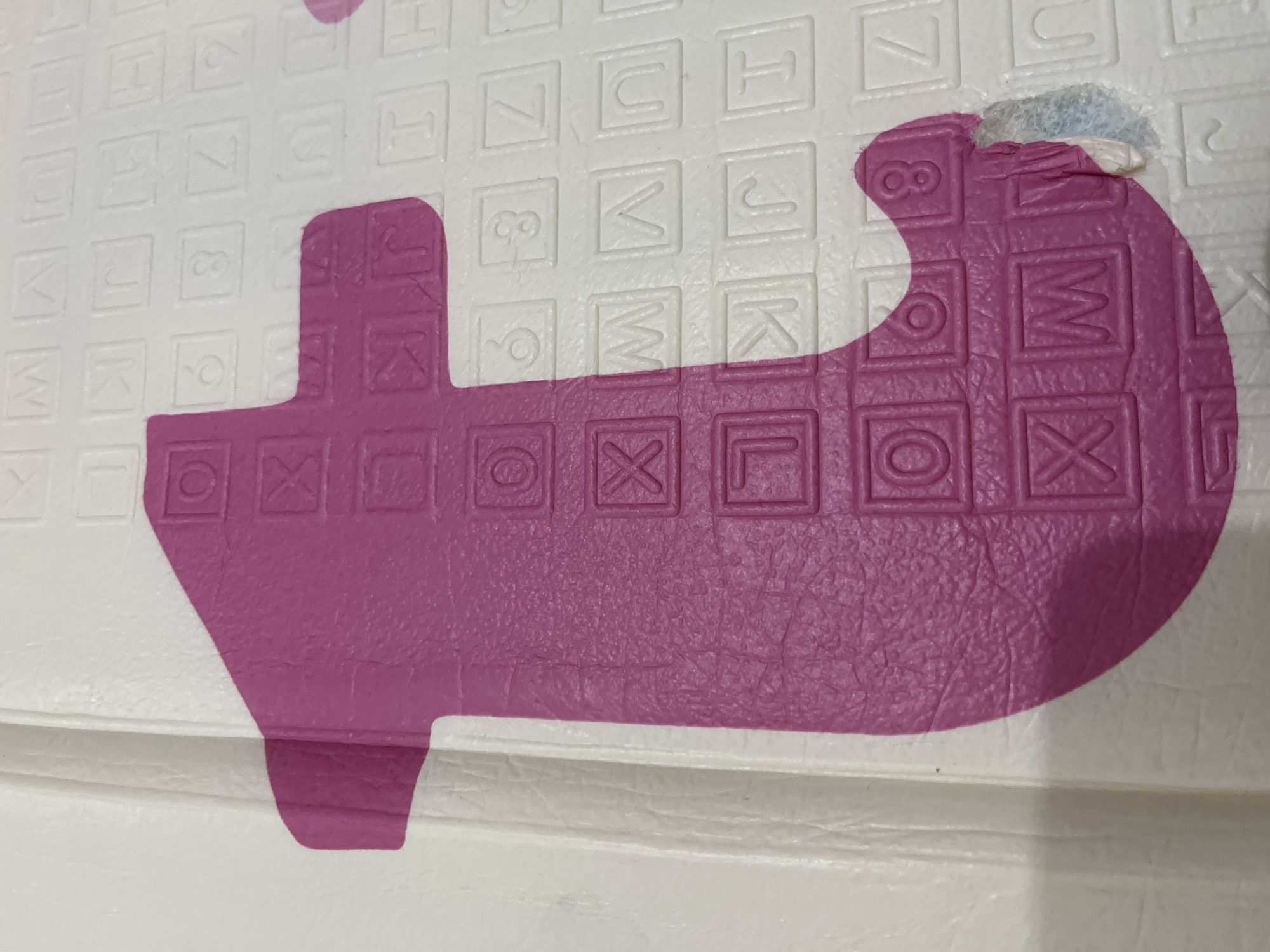 This image displays a close-up section of a children's foam puzzle playmat, situated in Lhokseumawe, Indonesia. The central subject is a large, magenta-pink, lowercase letter 't', characterized by a textured, slightly granular surface. This pink element is integrated into a larger white foam mat, which also exhibits a similar textured finish.
Embedded within the pink 't' are multiple debossed squares, each containing an uppercase letter or number in white, such as 'O', 'X', 'L', and 'M'. The surrounding white foam mat features numerous similar debossed squares, containing various uppercase letters and numbers, including 'W', 'V', 'U', 'K', 'J', 'I', '6', '3', '7', 'Z', '9', 'n', 'H', and 'T'.
At the bottom right curve of the pink 't', there is noticeable damage where the pink material is torn and peeled away, revealing an underlying lighter, possibly white, foam layer. The mat appears clean otherwise, with no other significant marks or debris visible. The lighting is uniform, highlighting the surface textures and the embossed details of the letters and numbers. No individuals, overt actions, or interactions are present in the frame.