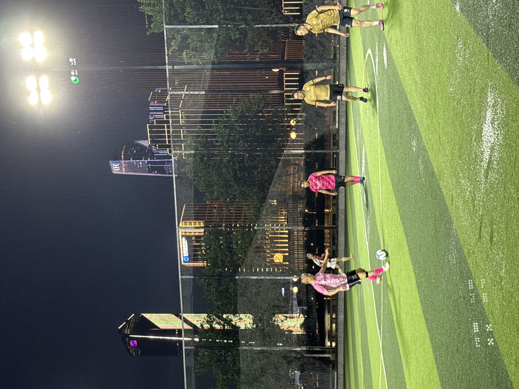 In Haizhu District, China, at night, several individuals are playing soccer on an illuminated artificial turf field, likely a sports complex or park, as indicated by the presence of floodlights. The backdrop features a cityscape with tall, modern buildings illuminated with colorful lights. Three individuals in yellow soccer jerseys and black shorts are on the field, along with two individuals in pink jerseys and black shorts. One person in a pink jersey is actively kicking a soccer ball. The scene is set against a dark sky, with visible light rain or water droplets in the air.