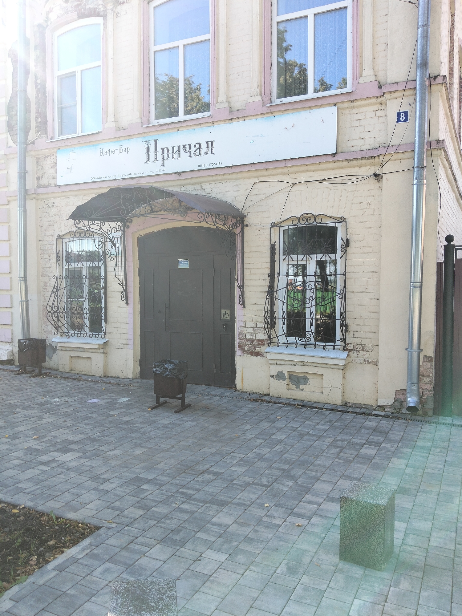 The image shows the facade of a cafe-bar named "Причал" (Prichal) located in Kozmodemyansk, Russia. The building is made of light-colored brick with pink accents around the windows and doorways. Above the entrance, a sign reads "Кафе-Бар Причал". The entrance consists of a large, dark wooden double door with decorative ironwork above it. To the left of the entrance is a window with a similar ornate iron grate. Two dark metal trash cans with black plastic liners are placed on the paved walkway in front of the building. The walkway is paved with grey rectangular tiles. To the right of the building, a tall metal downspout runs vertically along the wall, and a blue sign with the number "8" is visible on the wall. The overall scene depicts the exterior of a commercial establishment on a sunny day.