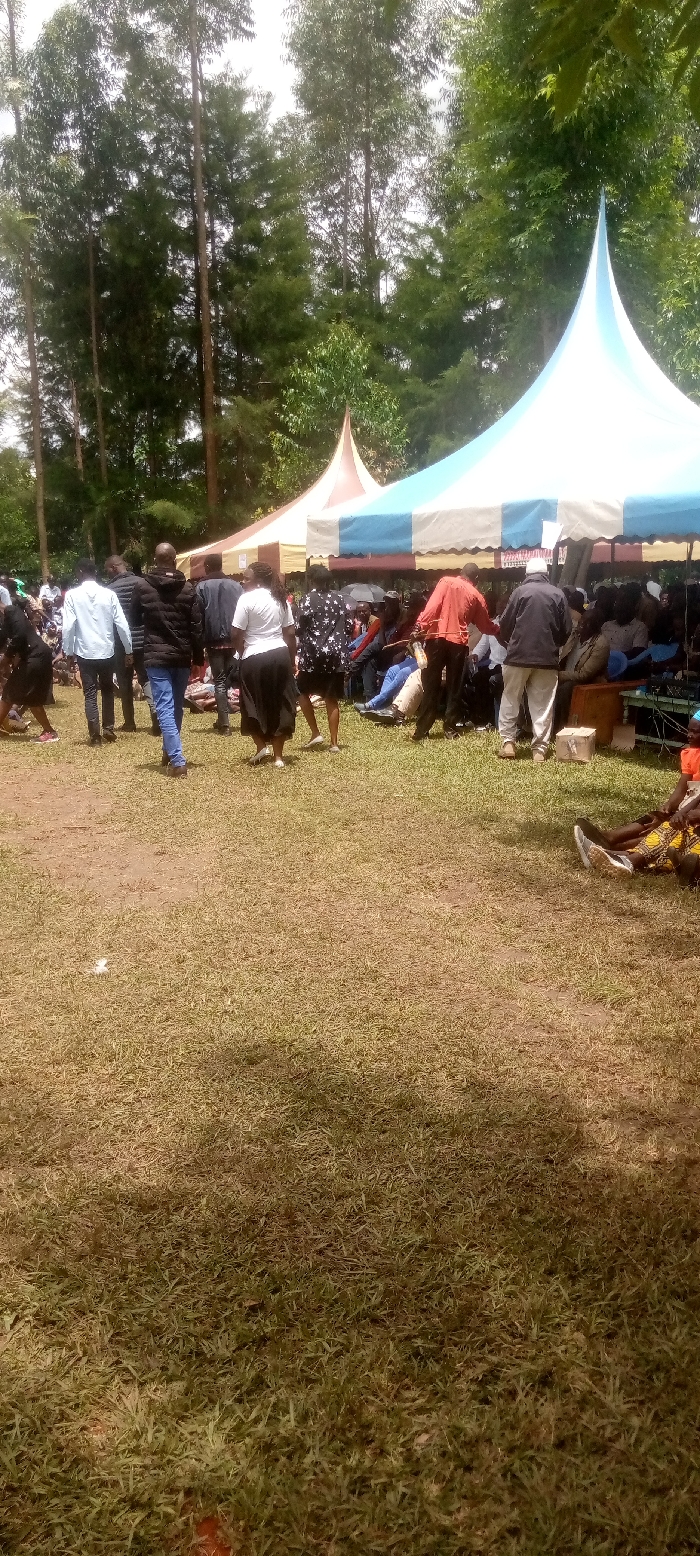 The image shows a group of people gathered in a grassy area under two tents, one blue and white and the other brown and white. The people are standing and sitting. The scene is a large grassy area with trees in the background. The location is near the coordinates of Burundu, Kenya, which is latitude 0.46, longitude 34.776. The image is taken from a low angle.