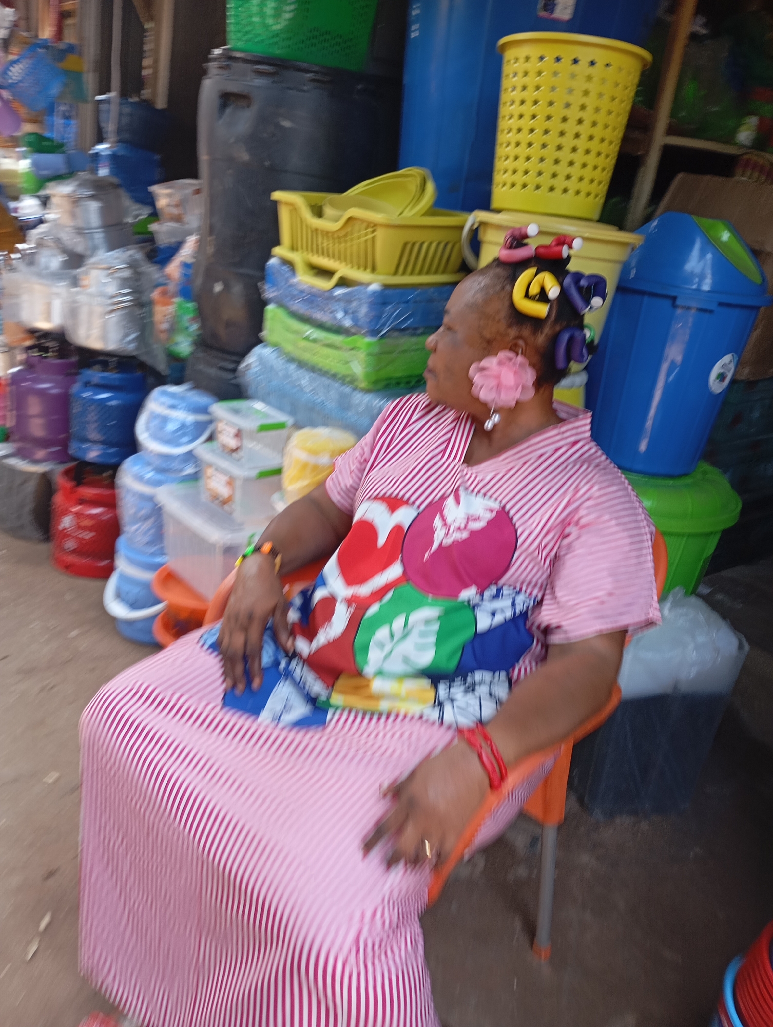 In Shendam, Nigeria, a woman sits on an orange chair in an outdoor market. She is wearing a pink and white striped dress with a colorful fabric draped over her lap. Her hair is styled with purple and yellow curlers, and she has a pink flower earring with a pearl. Behind her, various plastic containers and buckets are stacked, including blue, green, and yellow ones. A large blue trash bin is visible to her right, and further in the background, more market stalls and goods can be seen. The ground appears to be dirt.