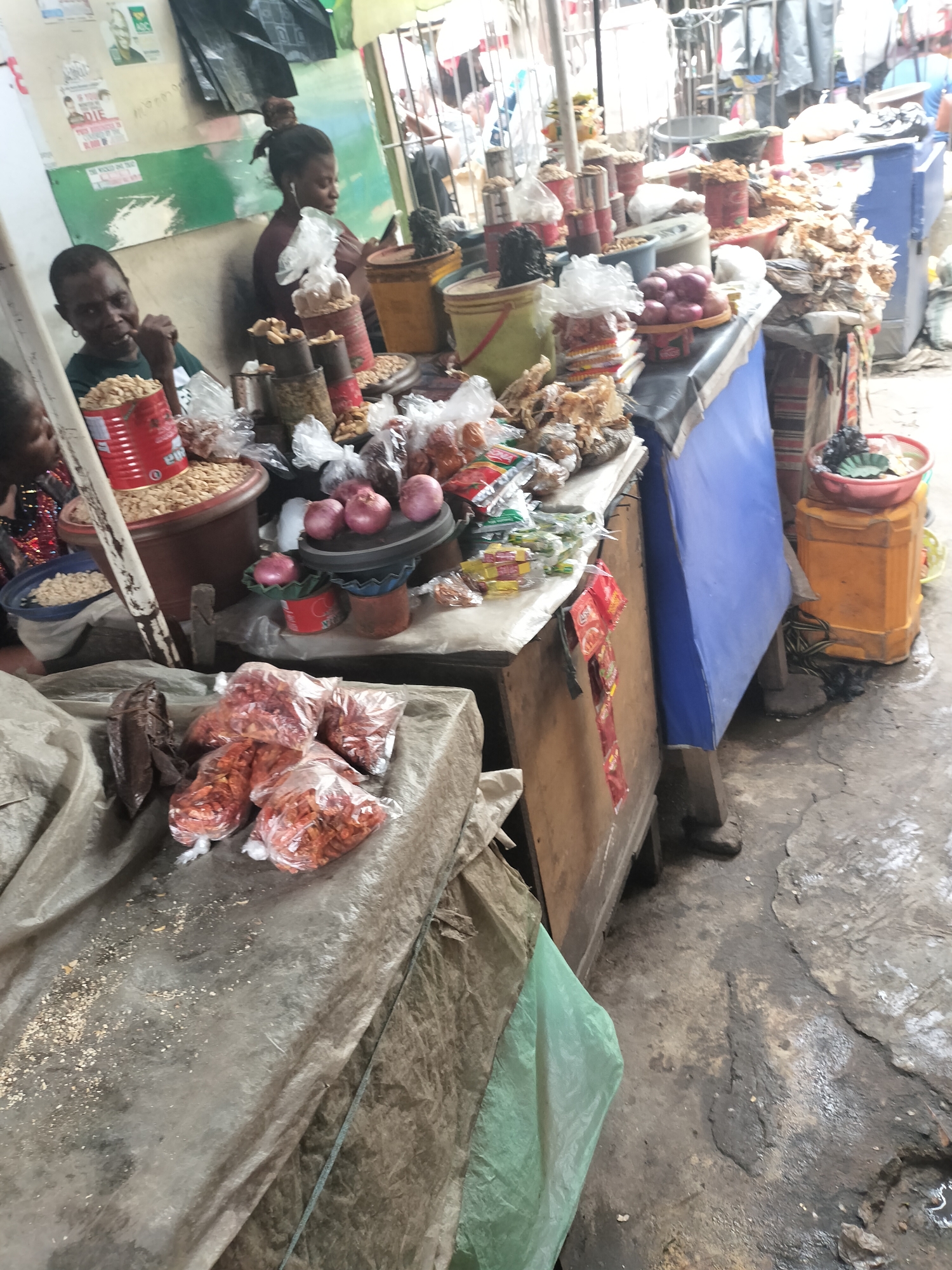 The image depicts a bustling market scene in Alaiyabiagba, Nigeria. Two women are seated behind stalls laden with various food items. One woman is observed eating, while the other is looking at a smartphone while wearing earphones. The stalls display a variety of goods including peanuts in a red tin, onions, various snacks in plastic bags, and other produce in containers. The environment is characterized by a somewhat crowded and gritty setting, with the ground appearing worn and stained. A blue tarp covers part of one of the stalls, and various buckets and containers are scattered around. Bags of what appear to be dried peppers are placed in the foreground.