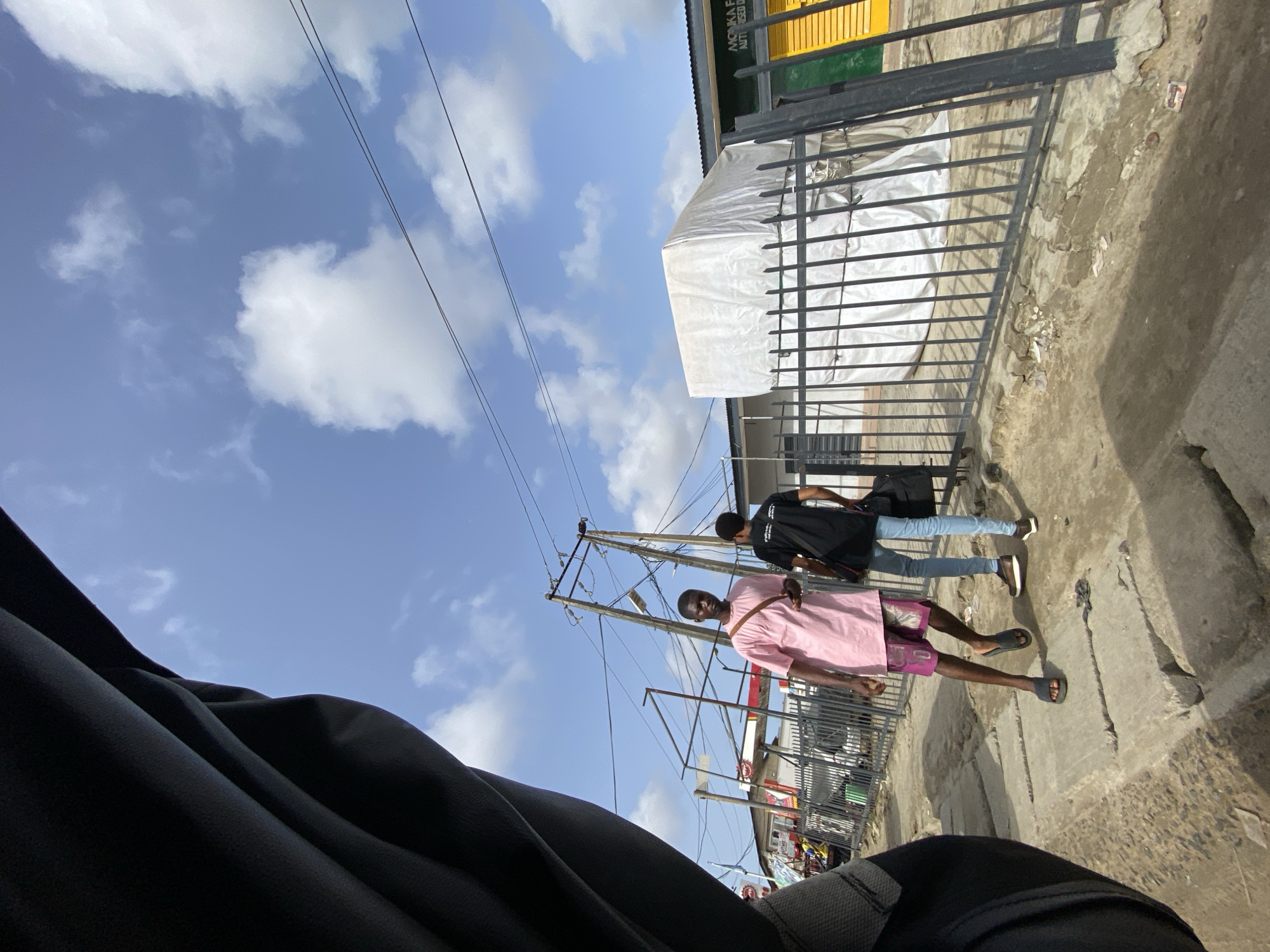 The scene is on a city street in Ado, Nigeria. Two young men are walking. The first man is wearing a pink top and purple shorts with sandals. The second man is wearing a black shirt and blue jeans. The background consists of a cloudy blue sky, buildings, power lines and poles, metal fencing, and an awning. The foreground includes a black material, possibly clothing. The street is concrete and appears worn.