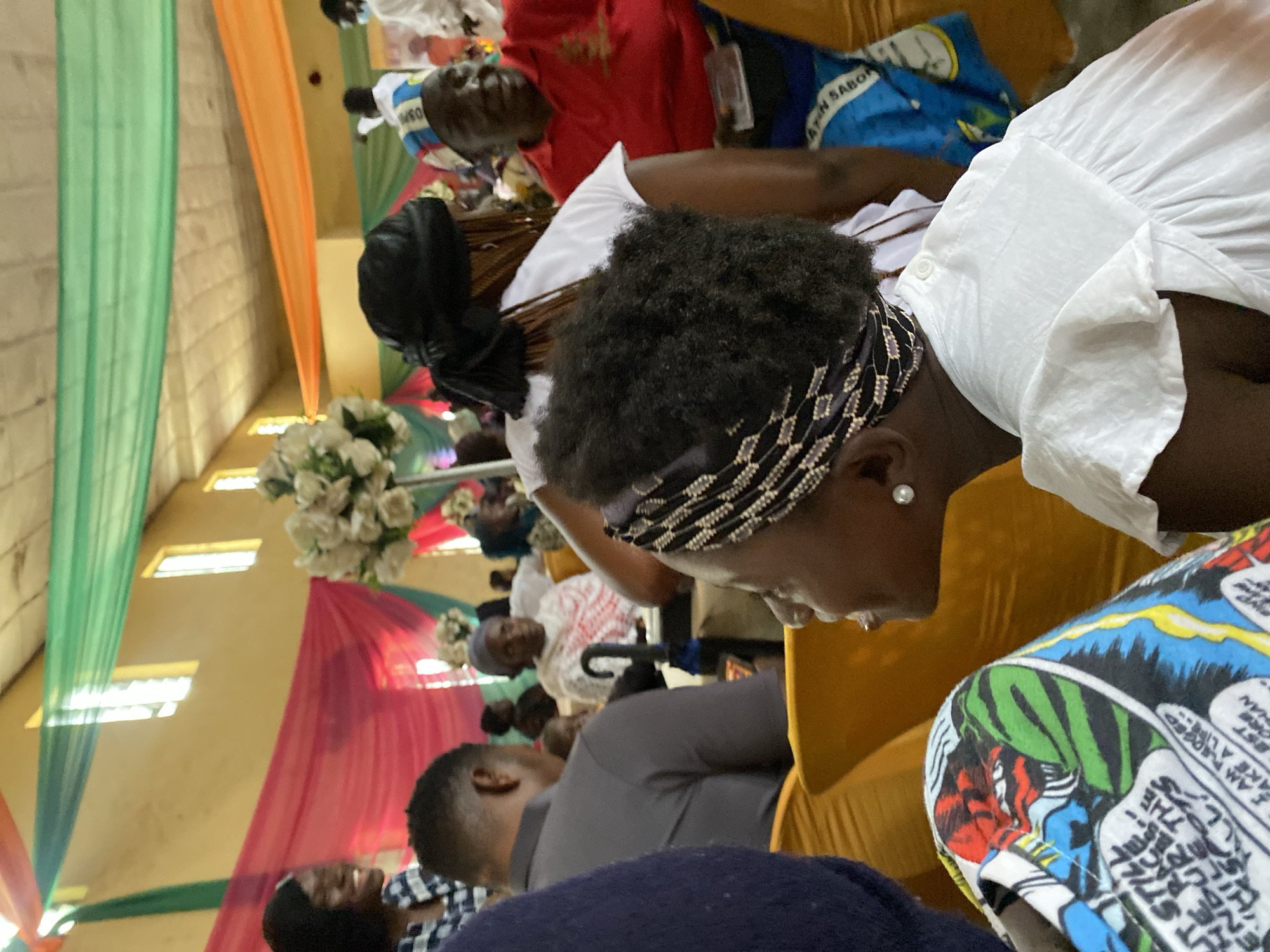 The image shows a gathering of people inside a hall decorated with draped fabric in shades of green, red, and orange. The hall appears to be a place of worship or a community event space. The lighting suggests it could be daytime, with natural light filtering in from windows visible at the top of the walls, or artificial lighting is in use.
In the foreground, a woman is visible from the side, wearing a white dress with ruffled sleeves and a black and white patterned headscarf. She has an earring in her visible ear. To her left, a portion of another person's lap is visible, covered by a colorful fabric featuring comic book style illustrations.
In the middle ground, several people are seated, some facing away from the camera and others in profile. A man in a grey shirt is seated to the left. Further back, a woman in a white outfit with red patterned fabric is seated, and to her right, a man in a red shirt and a blue and white patterned cap is looking towards the right.
In the background, more people are visible, some standing and some seated. A large floral arrangement of white flowers is placed on a stand in the mid-ground. The overall scene depicts a lively event or gathering. The location is identified as Turu, Nigeria.