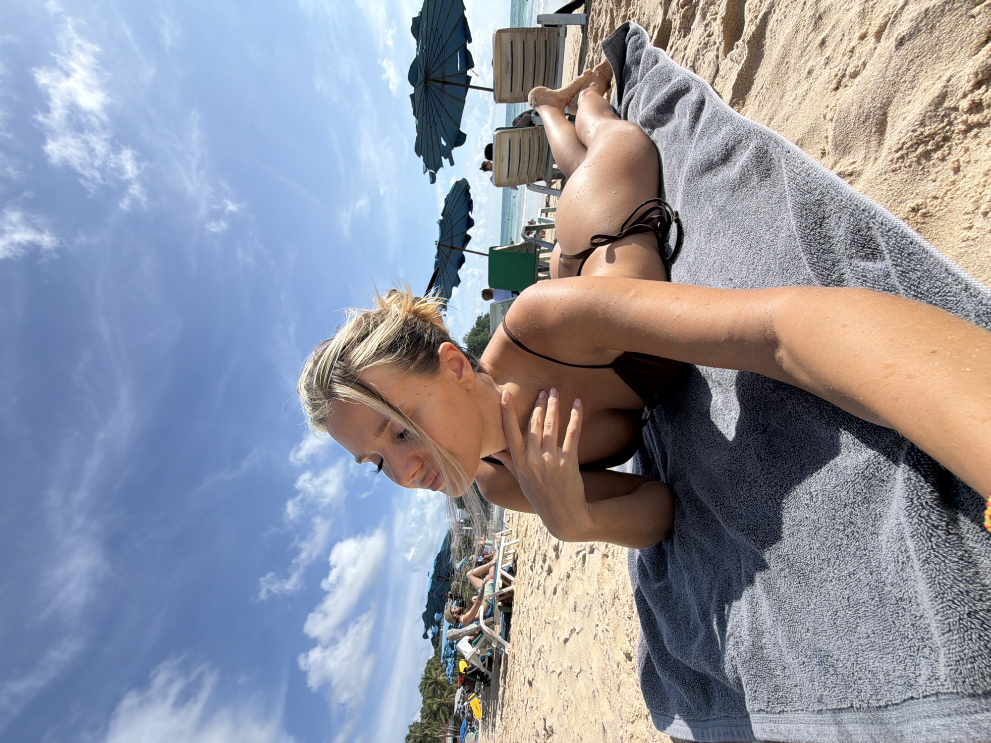A woman is lying on a beach towel on the sand at Karon Beach, Karon, Thailand. She is wearing a dark bikini and her hair is blonde and pulled back. She is lying on her stomach, with her head turned to the side and her eyes closed. Her right arm is resting on her neck. In the background, there are beach chairs, umbrellas, and other people. The sky is blue with some white clouds.