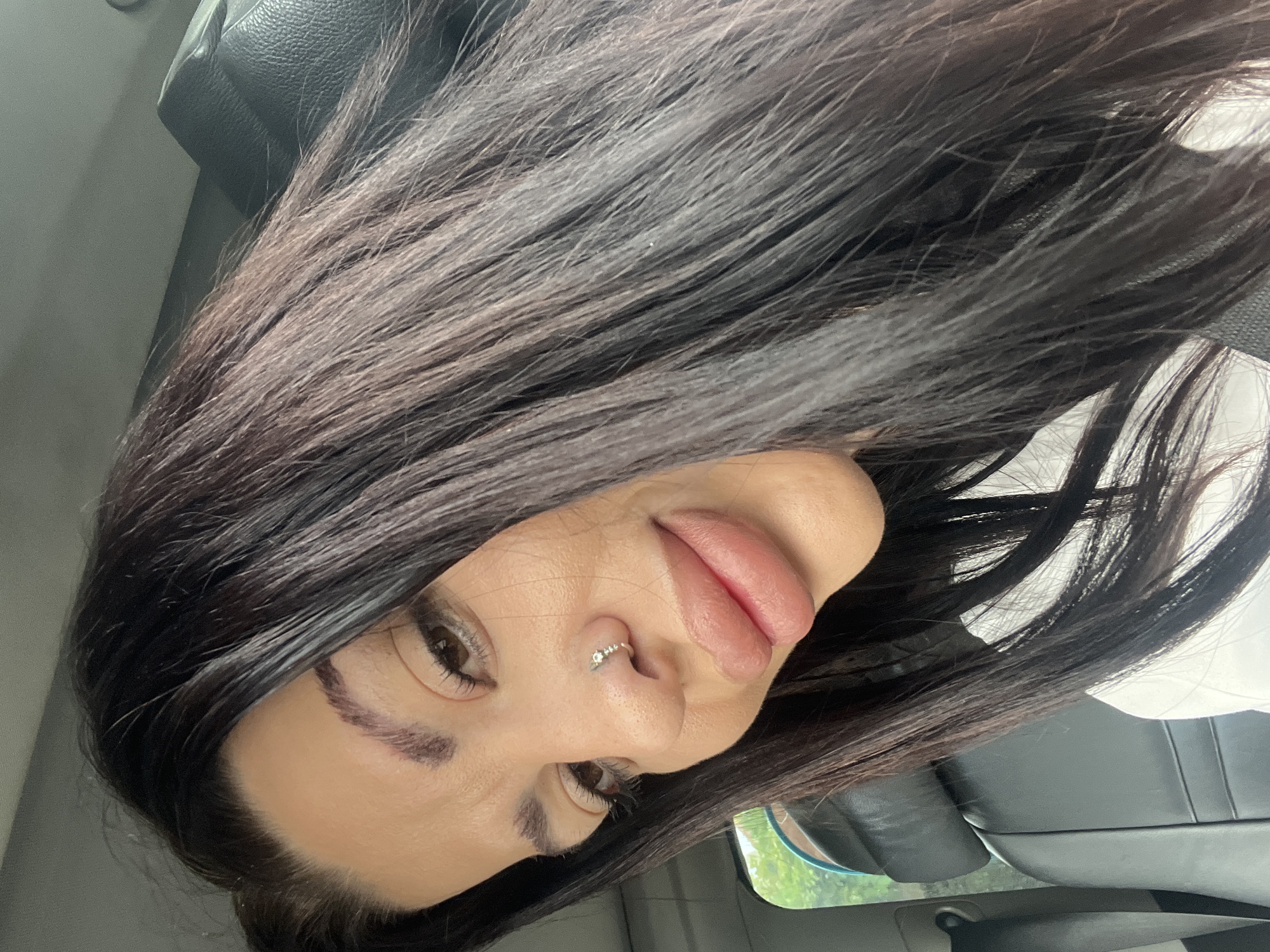 A close-up, high-angle, eye-level shot shows a woman with dark hair and fair skin looking at the camera. She is in the driver's seat of a car, with a black leather headrest visible behind her. The woman is wearing a white shirt. She has full lips and a small nose ring. The background is blurred, but a green outdoor scene is visible through the car window. The image was taken in Kalekovets, Bulgaria.