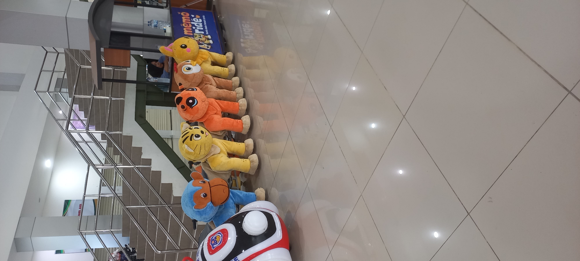 The image depicts a row of plush animal ride-on toys lined up on a tiled floor in an indoor public space, likely a mall or amusement center. The toys include a blue monkey, a yellow tiger, an orange creature resembling a tiger or lion, a brown bear, and a yellow rabbit. In the background, a staircase with stainless steel railings is visible, along with a kiosk or counter area. A person wearing a dark hijab is seated at the counter, and water bottles are displayed on a shelf behind them. A banner with the text "mémoride" and Indonesian text "Dunia main pental ceria" is also present. The lighting suggests an indoor environment, possibly with overhead lights. The floor is made of light-colored square tiles that reflect the overhead lights. The overall scene appears to be a place for children's entertainment.