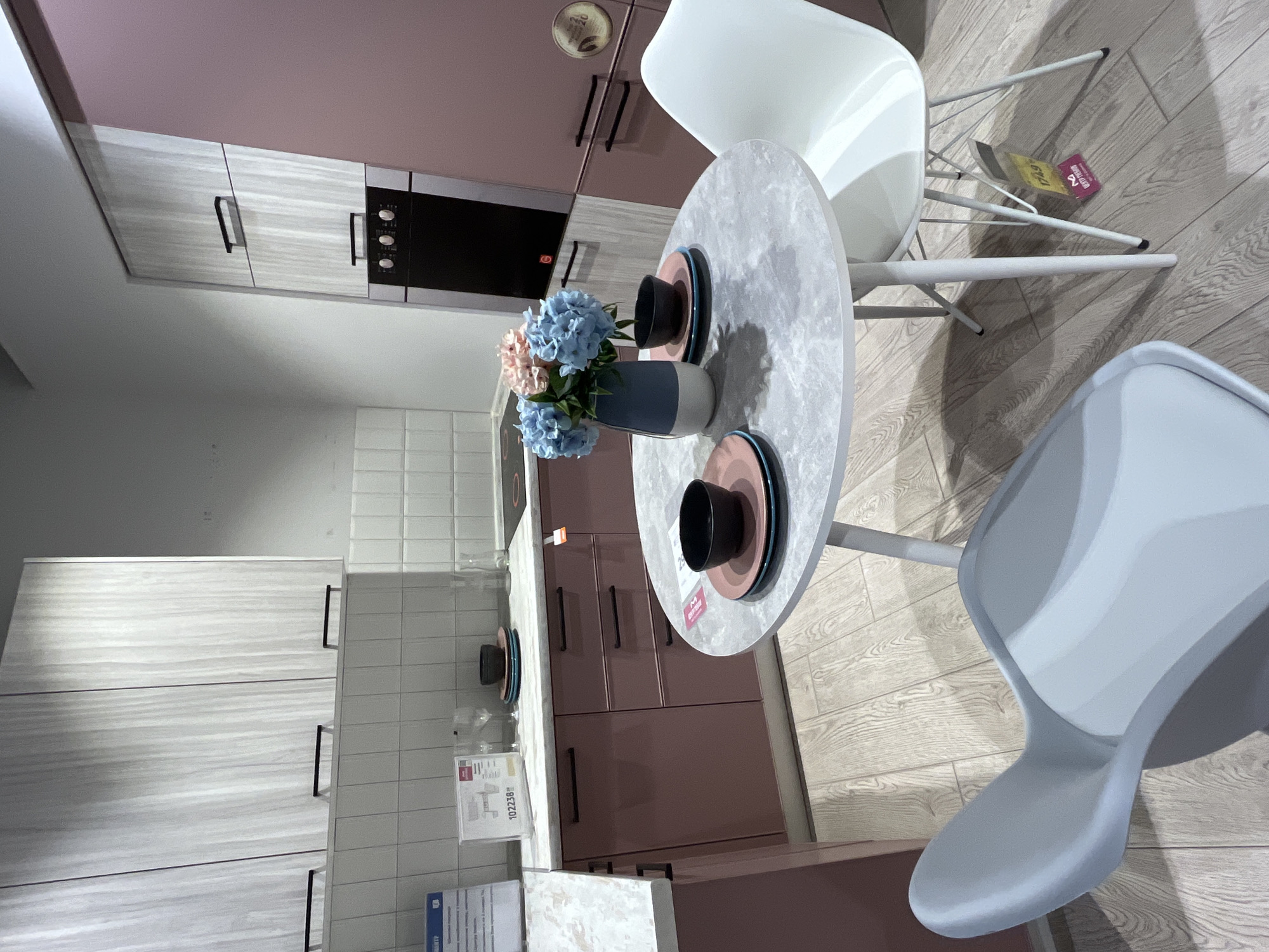This is a medium shot of a kitchen display in a store in Kharkiv, Ukraine. The kitchen features light wood-grain upper cabinets and muted pink lower cabinets and drawers with black handles. A white tile backsplash is visible above the countertops.
In the center of the image, a round, light gray speckled table stands on white legs. On the table, a dark blue and white vase holds a bouquet of blue and pink artificial flowers. There are two place settings on the table, each with a pink plate, a teal plate, and a black bowl.
To the right of the table, a white chair with thin metal legs is positioned. A yellow tag with red text is attached to one of the chair's legs. Another light gray chair with a molded seat is visible in the foreground to the left.
Behind the table, a black built-in oven with three knobs is integrated into a light wood-grain cabinet. The kitchen floor is made of light-colored wood-look laminate flooring. A countertop with a white, speckled surface runs along the back wall. A white sign with blue text is partially visible on the left side of the countertop.