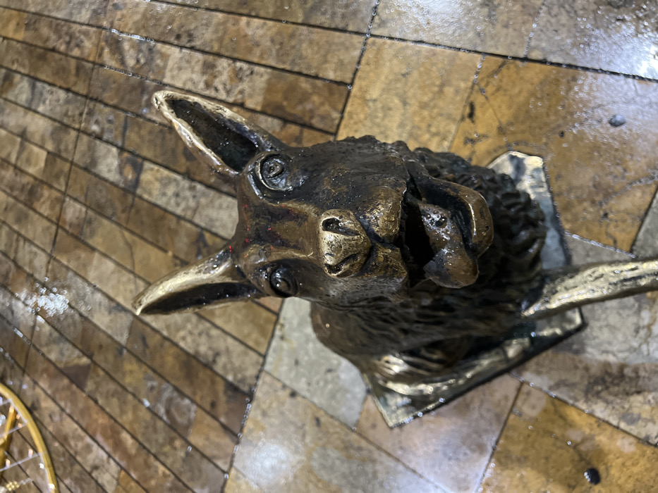A bronze or metal sculpture of a llama's head is mounted on a wall or plaque. The llama has detailed features like expressive eyes and textured fur. The metallic finish gives it an aged appearance. The background is a tiled floor or wall with a glossy surface, reflecting light and creating a wet appearance. The perspective is from above, looking down at the llama head.