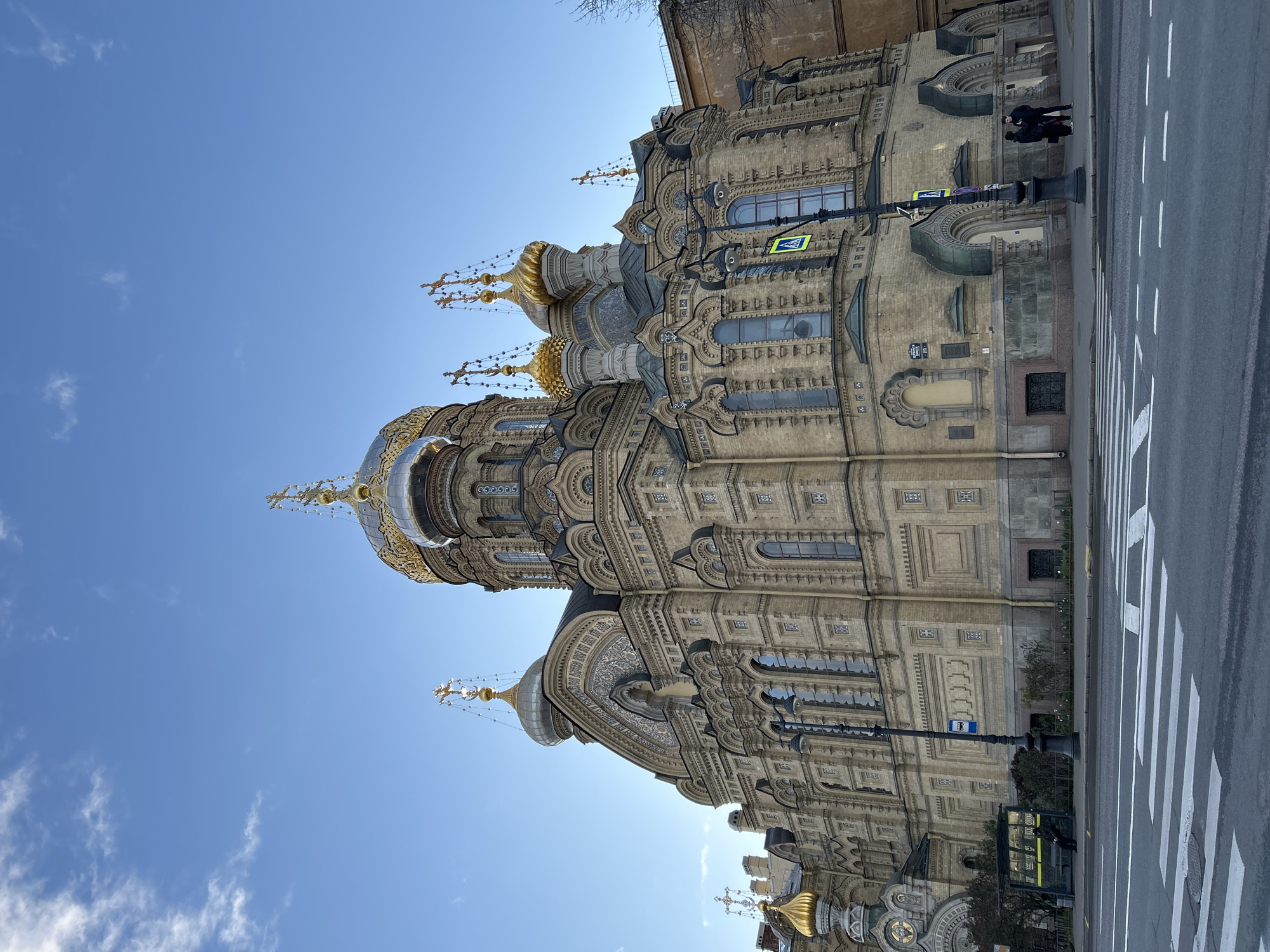 The image shows the facade and domes of the Church of the Savior on Spilled Blood in Saint Petersburg, Russia. The building is ornate and features multiple domes, each topped with a cross. The facade has numerous windows and decorative elements.
The photo is taken from a low angle, looking up at the church against a clear, blue sky with a few light clouds. Part of a crosswalk and a street are visible on the right side of the image, along with a few indistinct figures walking. A street sign and lamp post are also visible.