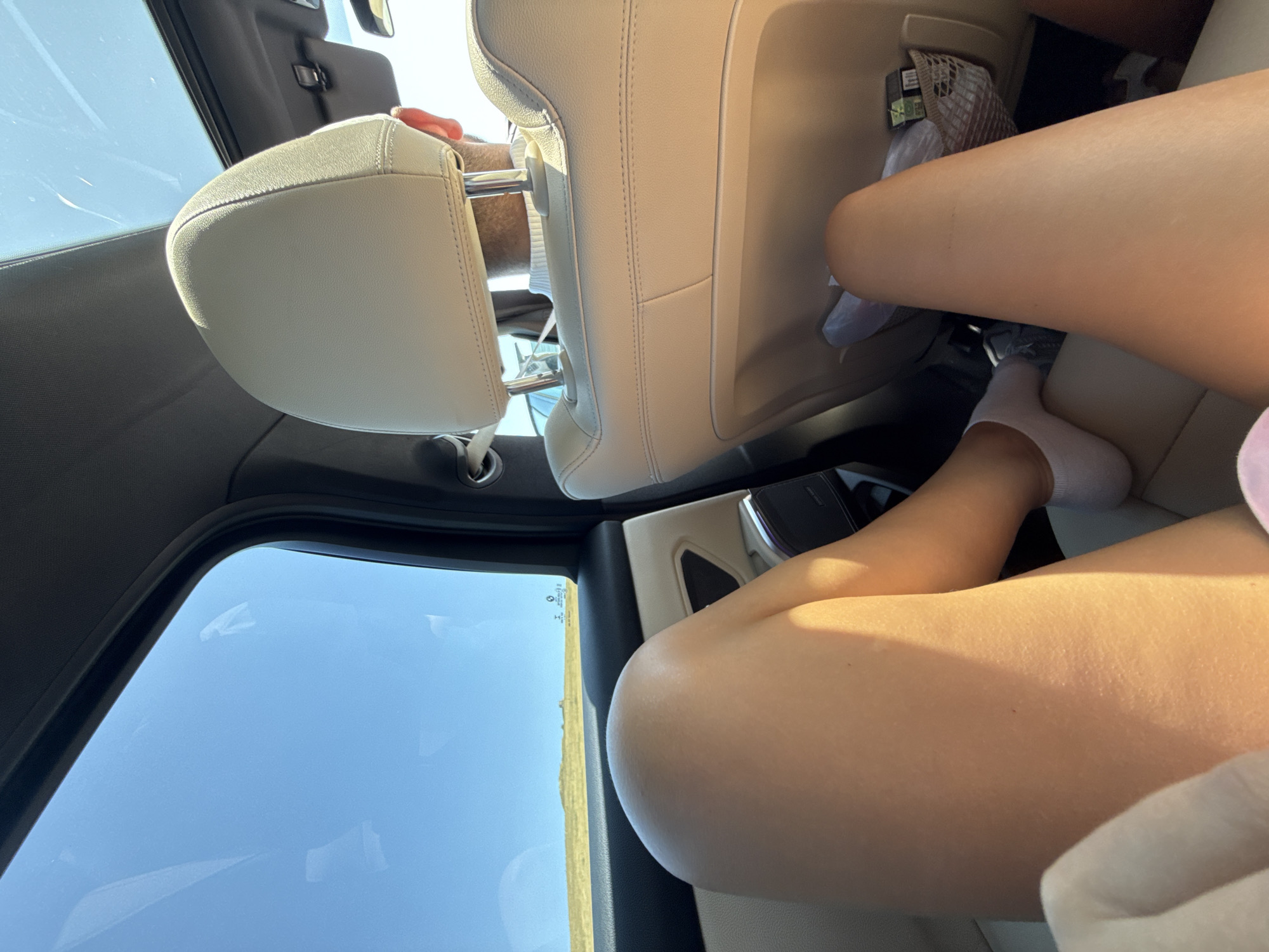 The image captures the interior of a car, likely a luxury model, with a focus on the rear passenger seats and the legs of individuals. The seating is upholstered in light-colored leather, with visible stitching. A panoramic sunroof extends over the rear of the car, revealing a bright blue sky. The scene appears to be a road trip, as indicated by the view of a landscape outside the window, suggesting a location outside of an urban environment. The landscape visible outside the window consists of rolling hills and sparse vegetation under a clear sky. A mesh pocket is attached to the back of the front passenger seat, containing what appears to be a small packet or package. One of the individuals in the rear has their legs prominently featured, wearing white socks. Another individual's head and shoulders are visible in the foreground, seen from behind, wearing a light-colored sweater. The overall lighting suggests it is daytime, with sunlight streaming in through the sunroof and windows, casting shadows and highlighting textures. The specific location within the city of Codlea, Romania, cannot be determined from the visual information provided.