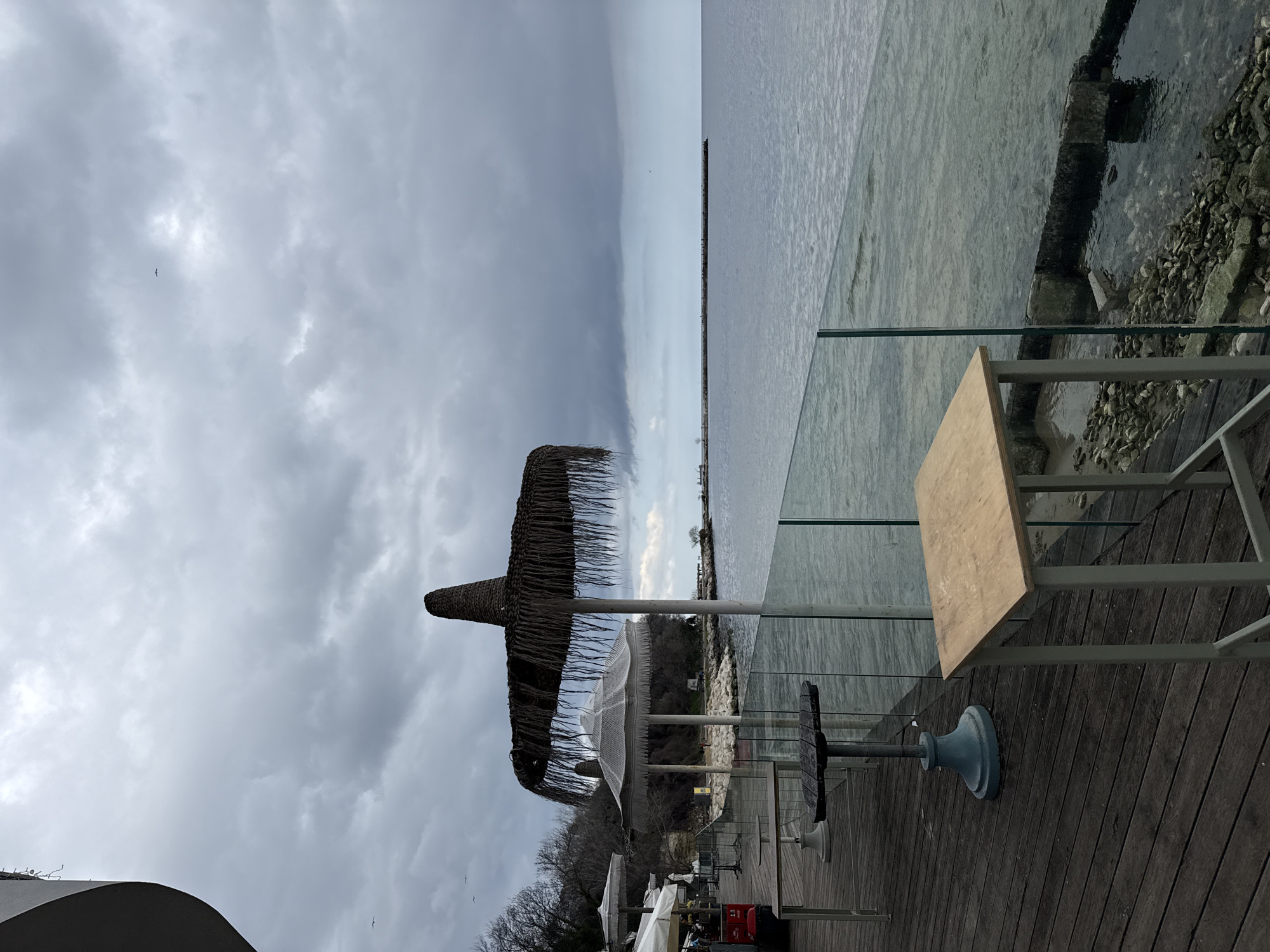 The image shows a seaside scene, likely a restaurant or cafe terrace, in Varna, Bulgaria. The foreground features a wooden deck with tables and chairs, some shaded by thatched umbrellas. A glass barrier separates the terrace from the sea, which appears calm under an overcast sky. A pier or breakwater stretches into the distance. The immediate shoreline consists of rocks and concrete structures. There are no people visible.