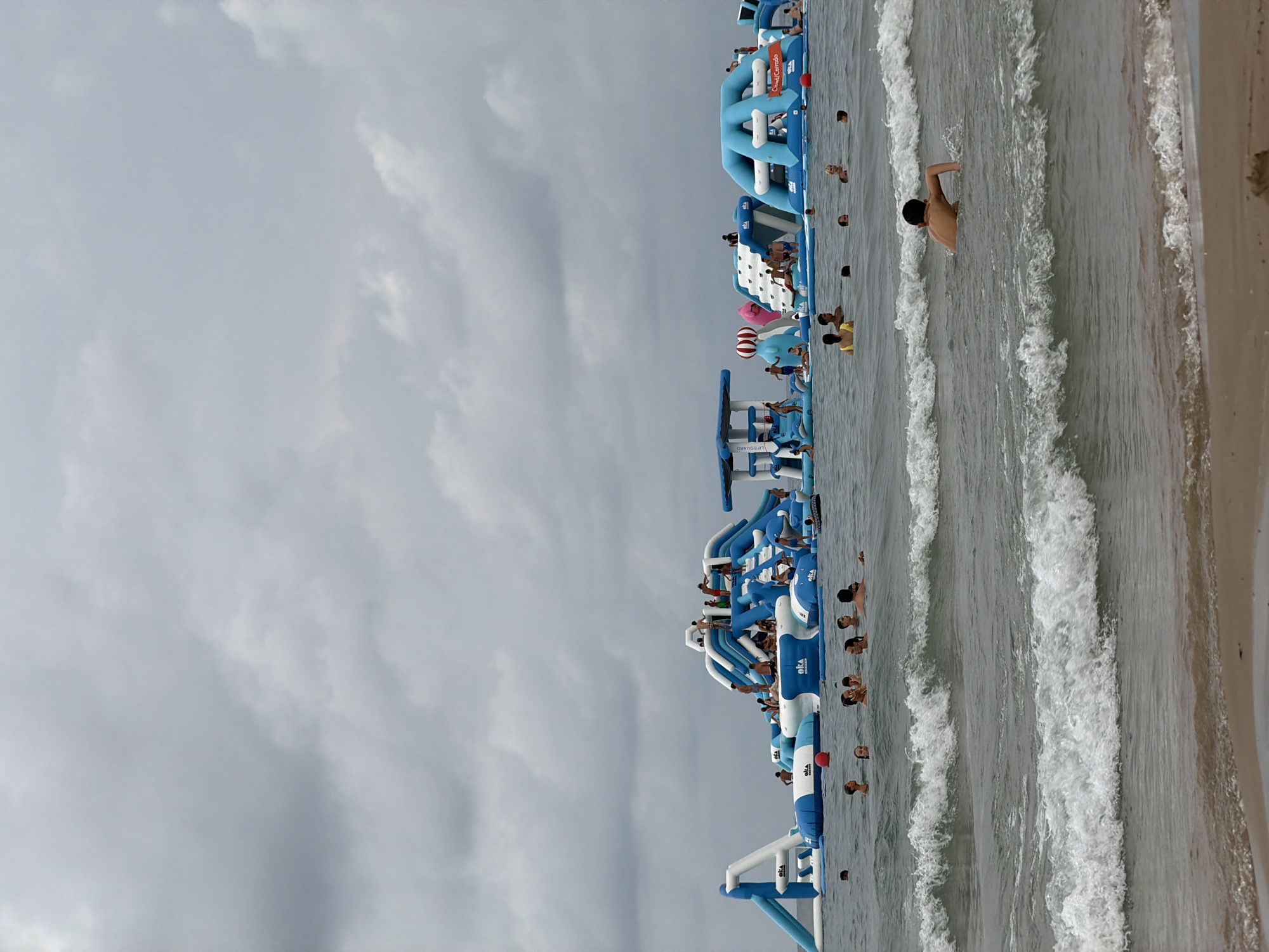 A wide shot captures an overcast day at an inflatable water park in Salou, Spain, situated in the Mediterranean Sea. Numerous people are actively engaged with the large, blue and white inflatable structures, which include slides, climbing walls, and obstacle courses. In the foreground, several individuals are swimming in the wavy water, with a young boy in the center appearing to be swimming towards the right. The water is greyish-blue with white-capped waves rolling towards a sandy beach. The sky is filled with heavy, grey clouds, indicating a potentially stormy or gloomy day. The inflatable structures dominate the mid-ground, with many people visible on and around them, enjoying the recreational facilities.