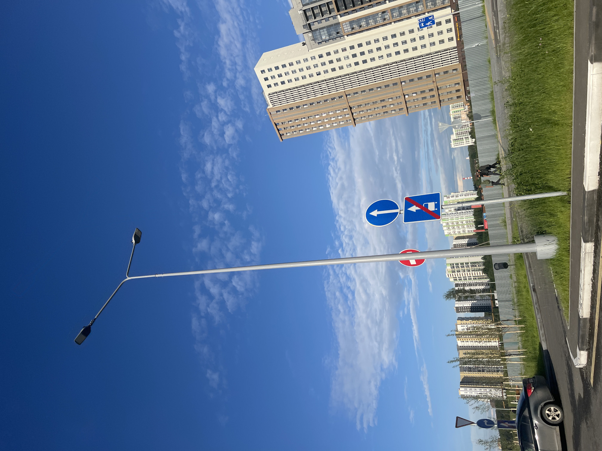 This image depicts a street scene in Yekaterinburg, Russia, under a bright blue sky with scattered white clouds. In the foreground, a gray sedan is parked on the left side of the frame, and a paved road curves towards the right. A tall, gray street lamp with two lamps at the top stands prominently in the center-left, casting a shadow on the road.
Several road signs are visible. One is a circular sign with a red border and a white horizontal bar, indicating "no entry" or "closed for all vehicles." Next to it, a rectangular blue sign displays a white upward arrow and a white icon of a car with a diagonal red line through it, signifying "no vehicles" or "forbidden for vehicles." Another blue sign further to the right shows a white icon of a bus stop.
In the mid-ground, a metal fence runs horizontally across the image, separating the road area from a grassy embankment. Behind the fence, a row of modern, multi-story residential buildings with varying architectural styles and color schemes are visible. The buildings are tall and appear to be part of a new development, with many windows visible on their facades. Some of these buildings are light-colored with brown and beige accents, while others are darker.
In the background, more buildings and structures can be seen, suggesting an urban or suburban environment. A few people, appearing as silhouettes, are walking on a path or sidewalk behind the fence. The overall environment suggests a clear day in a developing urban area.