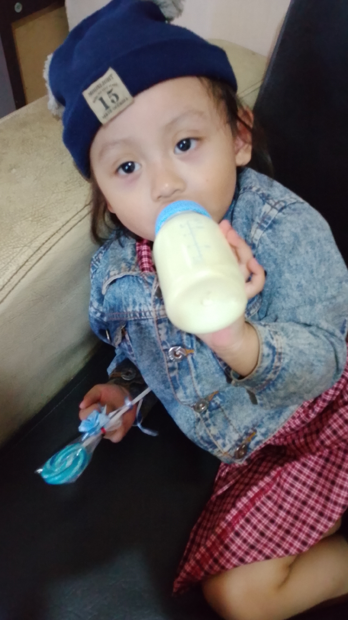 A young child wearing a blue knit hat and a denim jacket is sitting on a sofa and drinking from a baby bottle. The child also has a lollipop in their left hand. The background shows part of a sofa and a wall, suggesting an indoor setting. The image appears to be a personal photograph taken in Bojongsoang, Indonesia.