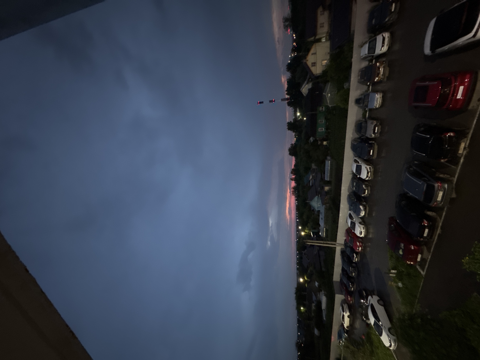 The image shows an aerial view of a parking lot filled with cars, situated in the village of Kondratovo, Russia. The scene is set during twilight, with a dark, cloudy sky. A faint pinkish hue is visible on the horizon, suggesting the sun has recently set. In the background, a cluster of houses with illuminated windows and streetlights can be seen, along with trees and a tall industrial structure with red and white lighting. The parking lot is paved and appears wet, possibly from recent rain. The cars are parked in rows, and include various models and colors, such as white, black, red, and grey. Some of the cars are sedans, while others appear to be SUVs or hatchbacks. The vantage point suggests the photo was taken from a higher elevation, possibly a building or balcony overlooking the parking lot and surrounding area.