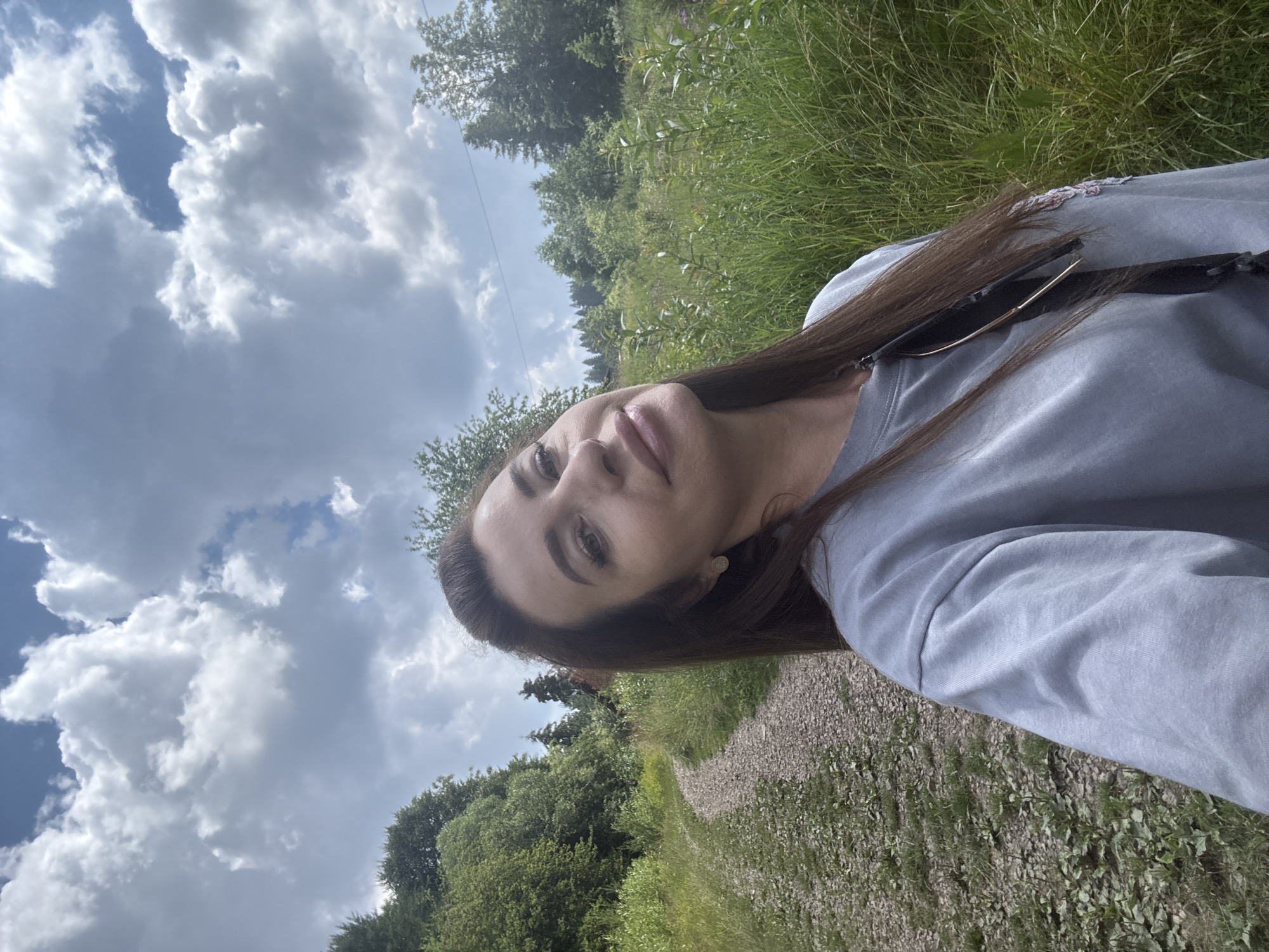 The image is a selfie taken outdoors on a cloudy day in Slavsko, Ukraine. The subject, a woman with long brown hair and fair skin, is positioned in the foreground, looking towards the camera. She is wearing a light grey long-sleeved shirt. The background shows a gravel path winding through lush green grass and trees. In the distance, there are more trees and a glimpse of what appears to be a building. The sky above is filled with dramatic, fluffy white clouds. The overall scene depicts a natural, rural environment.