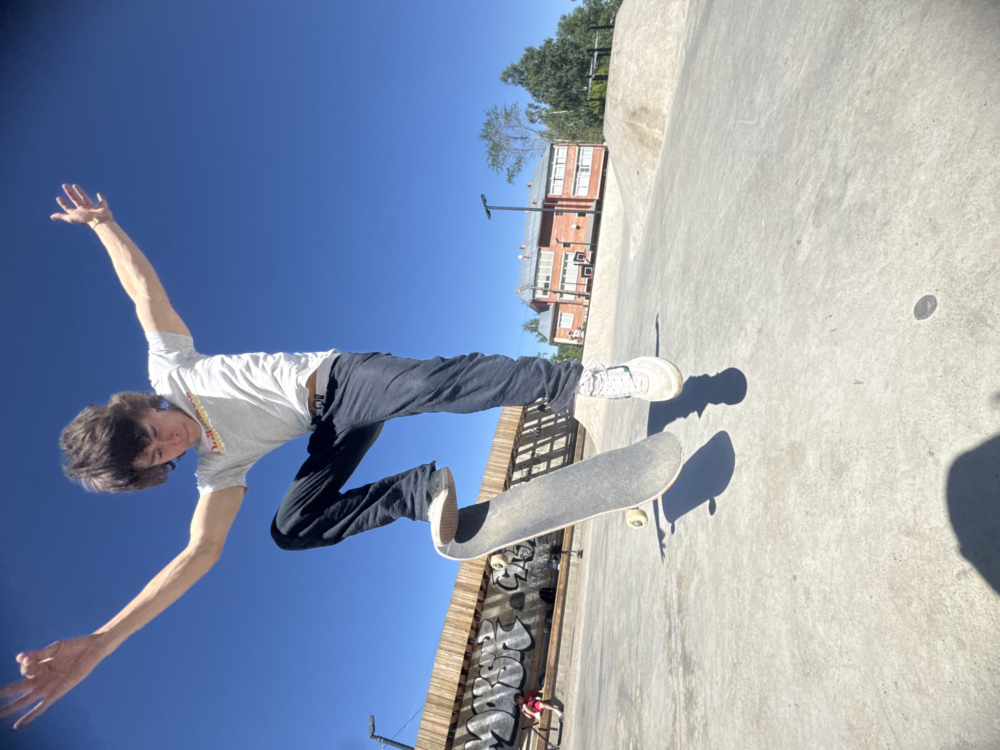 A young person with brown, shaggy hair is captured mid-air on a skateboard in a skate park under a clear blue sky. The person is wearing a light grey t-shirt with a colorful graphic, dark pants, and white sneakers. Their arms are extended outwards for balance, with the left hand positioned higher than the right. The skateboard is positioned beneath their feet, angled slightly upwards. The skate park features a concrete surface with ramps and sculpted mounds. In the background, there is a wooden fence with graffiti, a building with a red brick facade, and trees. The sunlight casts distinct shadows on the concrete ground. The overall scene depicts an action shot of skateboarding in an outdoor recreational area. This location is in the city of Divnogorsk, Russia.
