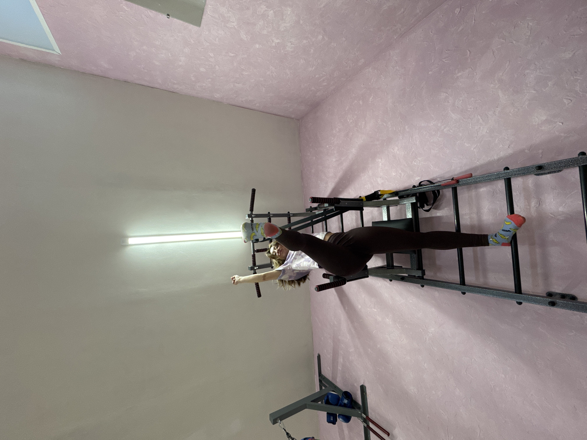 A woman performs a split on a wall-mounted Swedish ladder inside a room with textured pink walls and a white ceiling in Прикамский, Russia. The ladder is made of gray metal. The woman is wearing brown leggings, a light-colored top, and socks with a banana pattern. Her left leg is extended along the ladder, while her right leg extends in the opposite direction. One arm is raised above her head. Below the ladder is a pull-up bar.