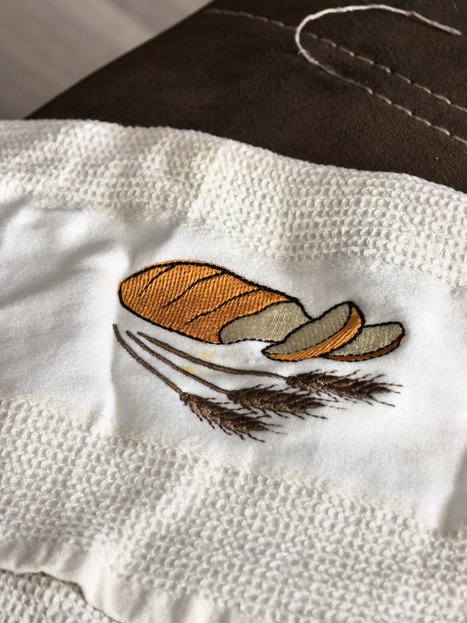 The image shows an embroidered image of a loaf of bread with slices and wheat stalks on a white towel. The towel appears to be on a brown surface, possibly a piece of furniture. There are no people or actions depicted in the image. The image does not contain any information that would allow for its location in Sterlitamak, Russia to be determined.