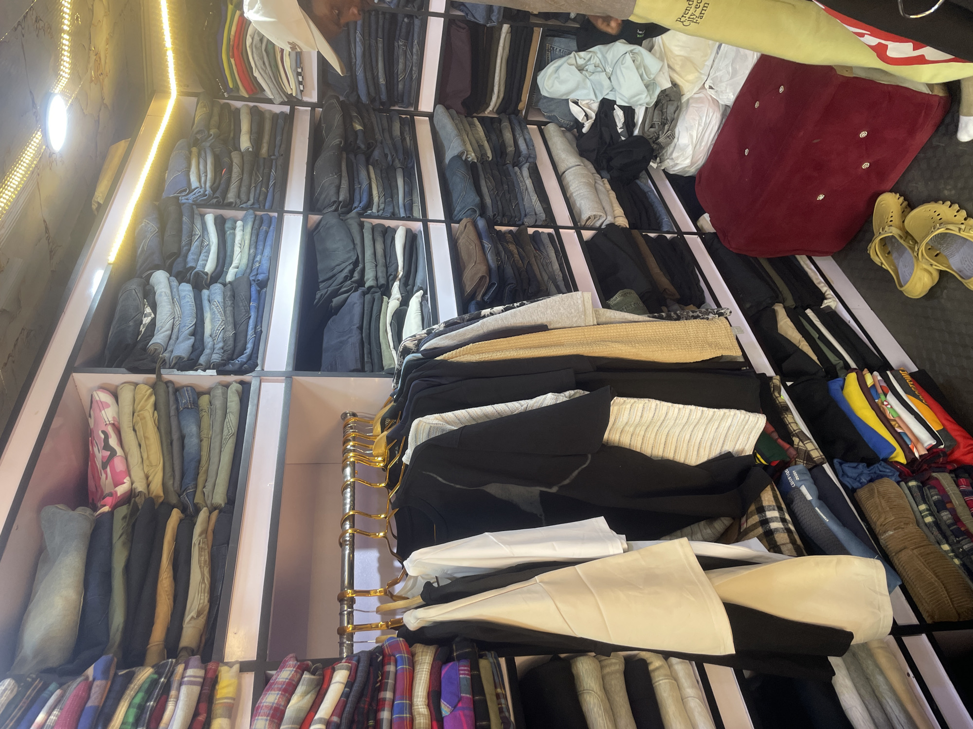 The image shows a cluttered retail space. Racks and shelves are filled with folded and hanging clothing items, including jeans, shirts, and jackets. The shelves are painted in a light pink color. The floor contains additional items, such as a red cushion, shoes, and bags. There is a person wearing a white hat visible in the upper part of the image. The location is stated to be Jos, Nigeria.