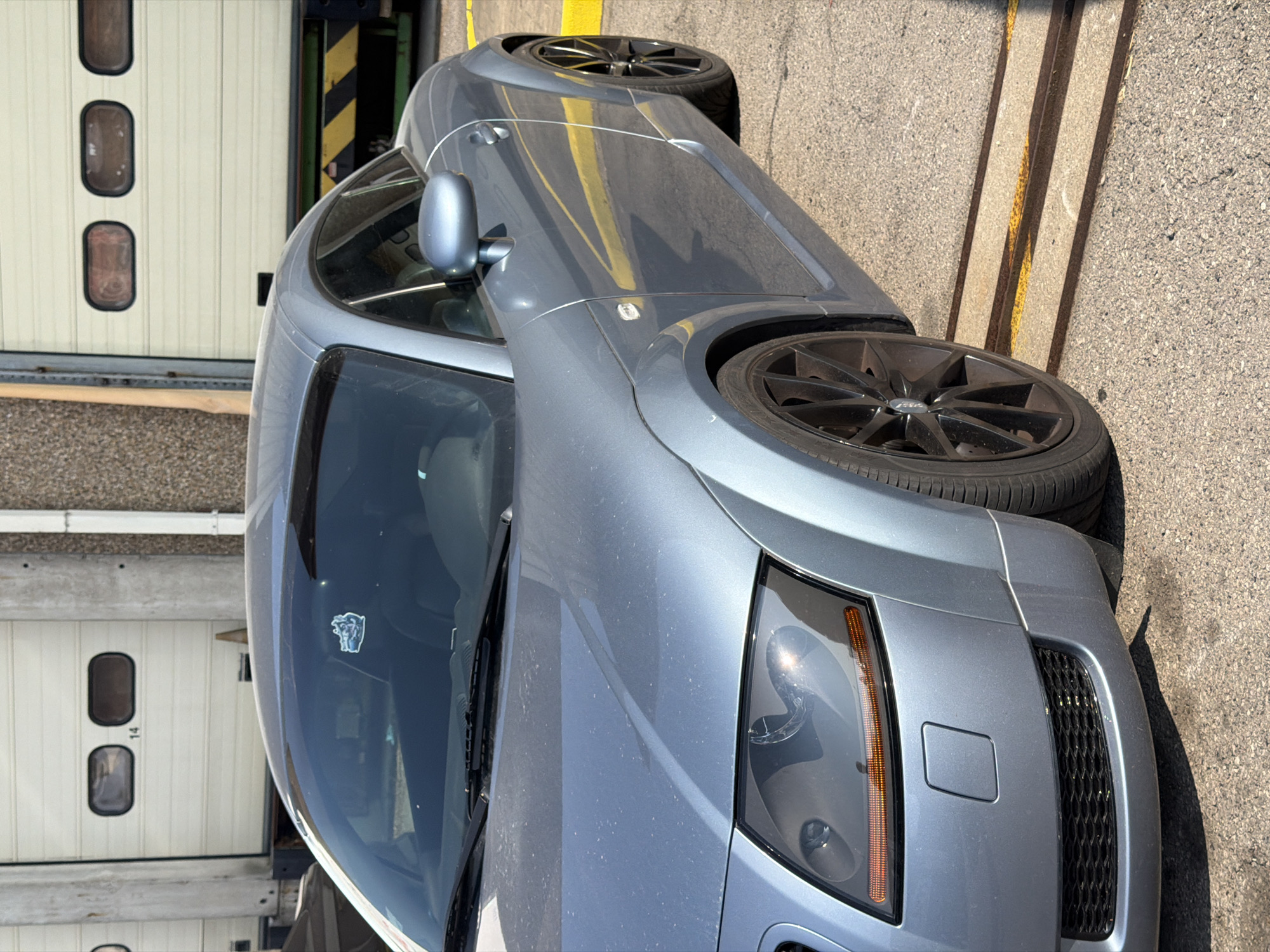 The image shows a close-up, eye-level view of a blue Audi TT parked on asphalt, with a segment of a large industrial garage door in the background. The garage door is white with horizontal panels and features two oval windows on the left and two on the right, with the number "14" visible between two windows on the left. A yellow and black striped barrier is visible at the bottom right of the garage door opening. The car's hood, windshield, driver's side door, front fender, and front wheel are prominently featured. A small sticker is visible on the interior passenger side of the windshield. The ground is asphalt with some visible gravel. The overall scene suggests an industrial or commercial area, possibly a depot or warehouse. The location is identified as Leudelange, Luxembourg.