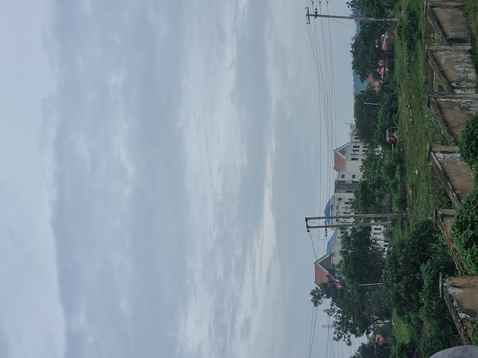 The image depicts a hazy, overcast sky over a semi-urban landscape, possibly in Rafin Par, Nigeria. In the middle ground, a cluster of multi-story buildings with white facades and various roof colors, including orange and blue, are visible amongst lush green trees. Some of these buildings appear to be under construction or recently built, with visible windows and architectural features. Electrical poles with wires crisscross the scene, indicating utility infrastructure. To the left, a small, red and yellow auto-rickshaw is parked in a grassy area. The foreground is dominated by a weathered, low-lying concrete structure with a stepped top, overgrown with green vegetation. The overall impression is of a developing area with a mix of residential buildings and natural greenery under a diffused light.