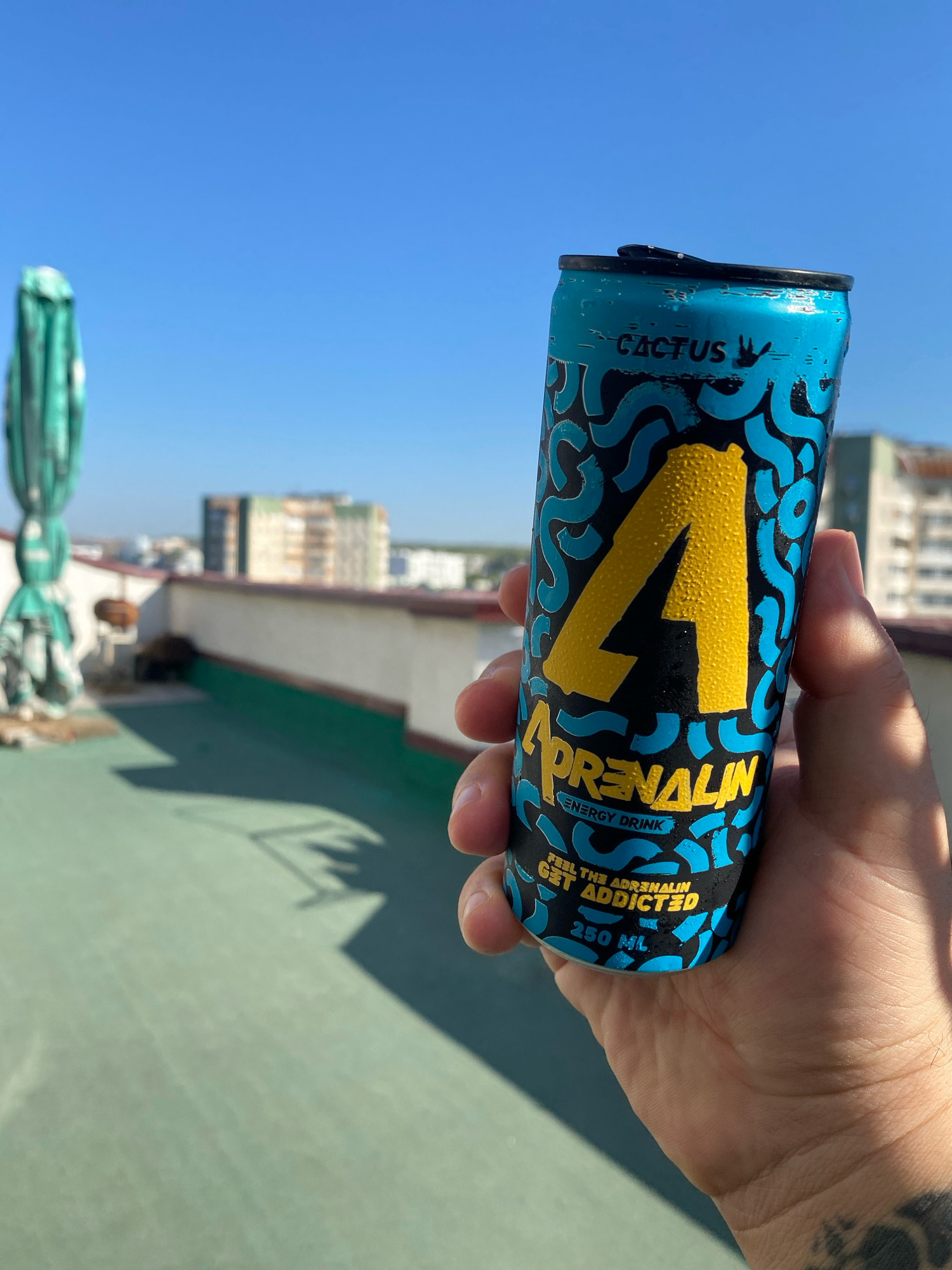 A person holds a blue and yellow can of "Adrenalin Energy Drink" with a cactus flavor. The can has a "4" prominently displayed on it. The background shows a city skyline with buildings, a green rooftop, and a clear blue sky. The person's hand holding the can shows some tattoos. The location is Chișinău, Moldova, on a rooftop with a view of apartment buildings.