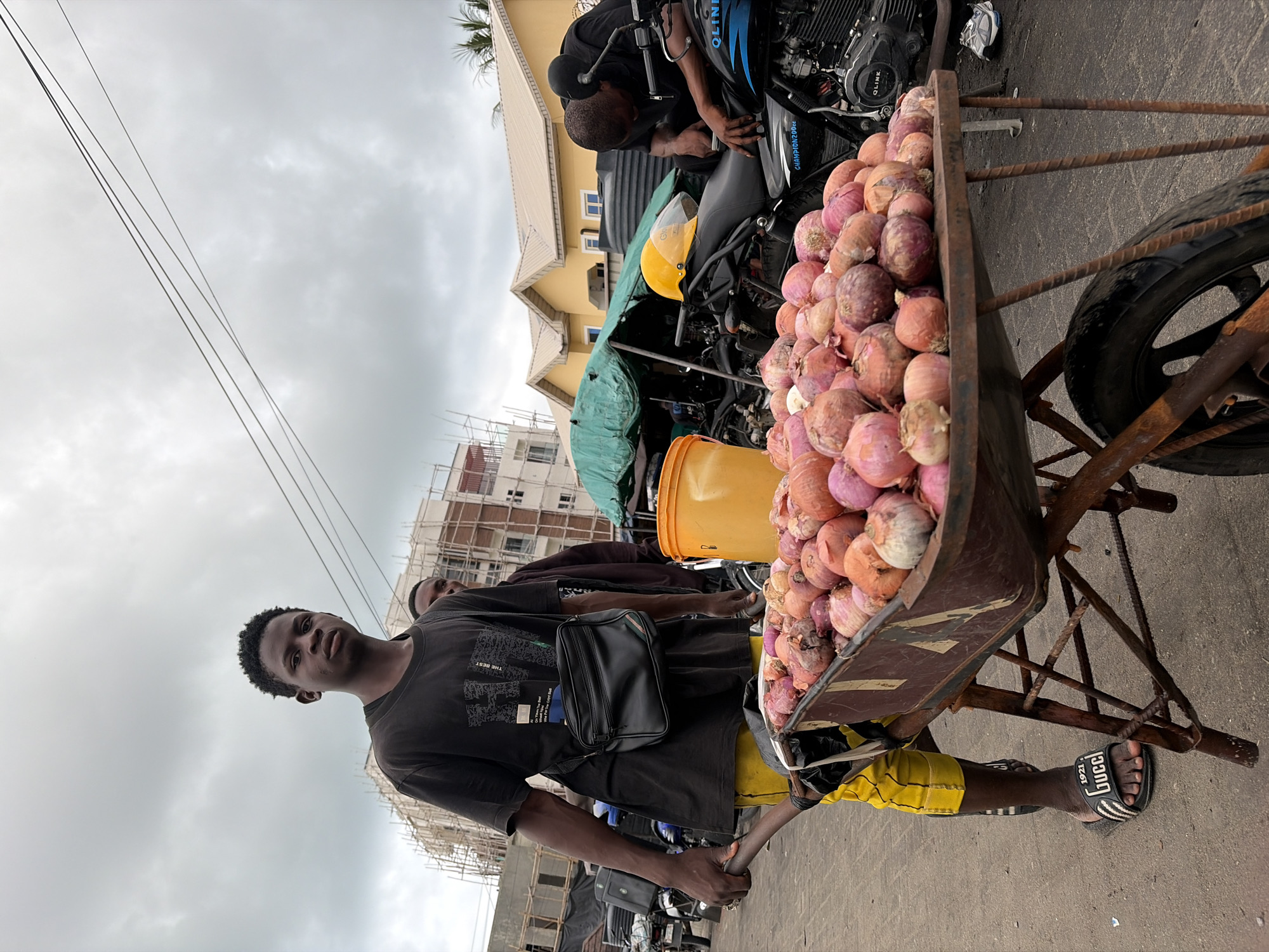This outdoor photograph, taken in Igboefon, Nigeria, captures a street scene under an overcast sky. In the foreground on the left, a young man with dark skin and short dark hair looks directly at the camera with a neutral expression. He is wearing a black t-shirt with "THE BEST" printed in white text, yellow and black patterned shorts, and black slides that display "Gucci 1921" on the strap. A black cross-body bag is worn across his chest as he holds the handle of a rusty metal wheelbarrow.
The wheelbarrow, prominently positioned on the right side of the frame, is heavily laden with numerous red and purple onions. Behind it, in the mid-ground, another man is bent over a dark motorcycle, seemingly engaged in maintenance. The motorcycle has "QLINK" and "CHAMPION 200cc" visible on its body, and a yellow helmet rests on its seat. An orange and a yellow plastic bucket are placed nearby.
The background features a light yellow house with a distinct roofline and blue-framed windows. Further back, unfinished buildings with scaffolding suggest ongoing construction or development. Thin power lines traverse the grey, cloudy sky above. The ground appears to be paved. The scene depicts everyday street commerce and activity.