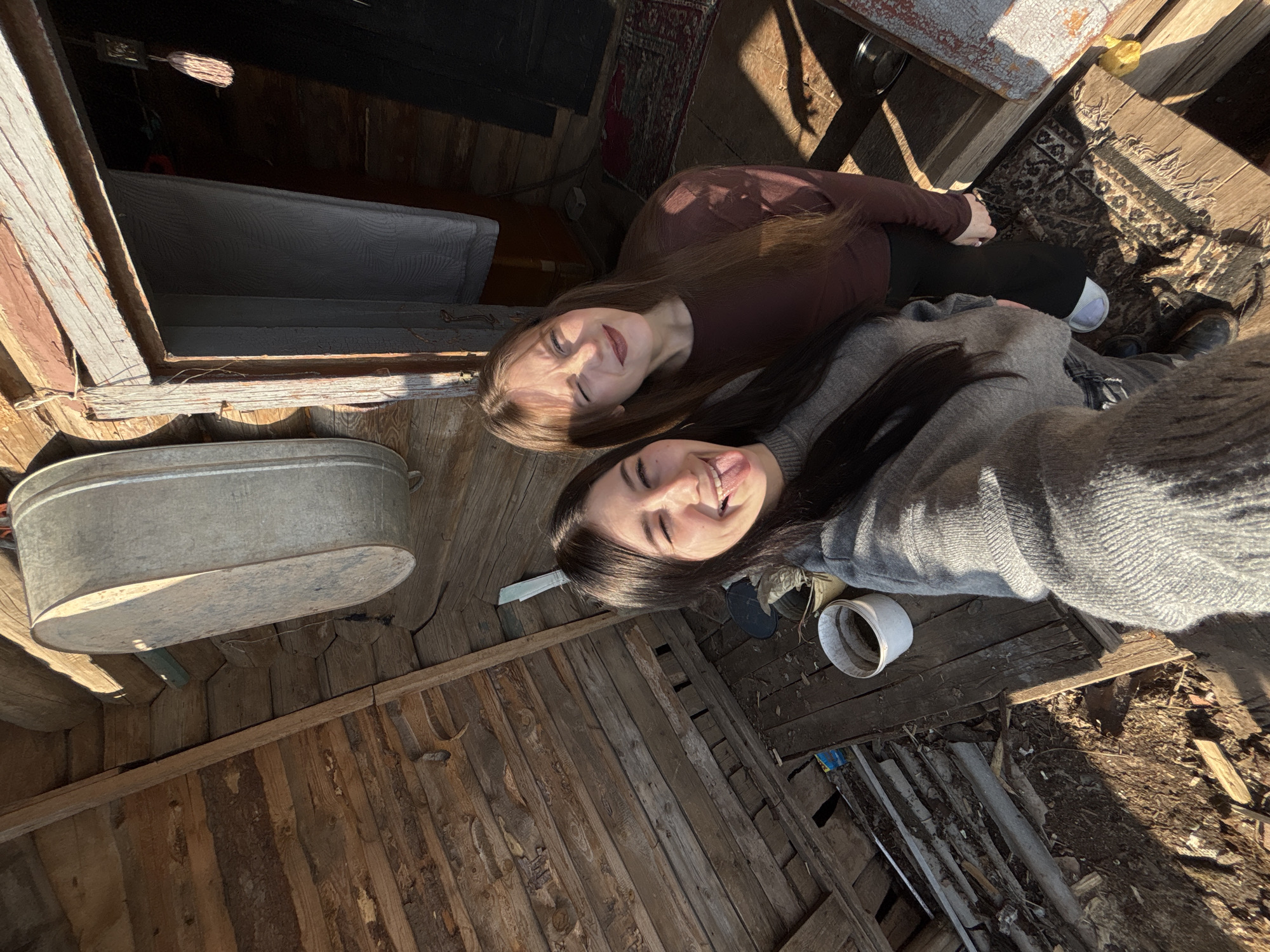 Two young women are depicted in a selfie taken in the city of Фоки, Russia, within a rustic, partially enclosed wooden structure. The woman on the left, with long dark hair, wears a gray knitted sweater and jeans; she winks her right eye and extends her tongue playfully while holding the camera. The woman on the right, with long light brown hair, wears a dark reddish-brown long-sleeved top and dark pants; she also winks her right eye and smiles. They stand on a wooden plank floor, with a section of disturbed earth and debris visible to the left, including a white plastic bucket. To the immediate right, an open doorway with a dark red frame leads into a darker interior, partially covered by a light-colored fabric. A small, patterned rug lies on the wooden floor near the doorway. Strong direct sunlight illuminates the scene, casting distinct shadows. A large, oval-shaped galvanized metal tub hangs vertically on the left wooden log wall. Exposed wooden logs and planks form the walls and overhead structure. A metal sickle is partially visible hanging on the upper left wall. A dark metal bowl rests on the floor to the right of the second woman's feet, and a small yellow object is visible nearby.