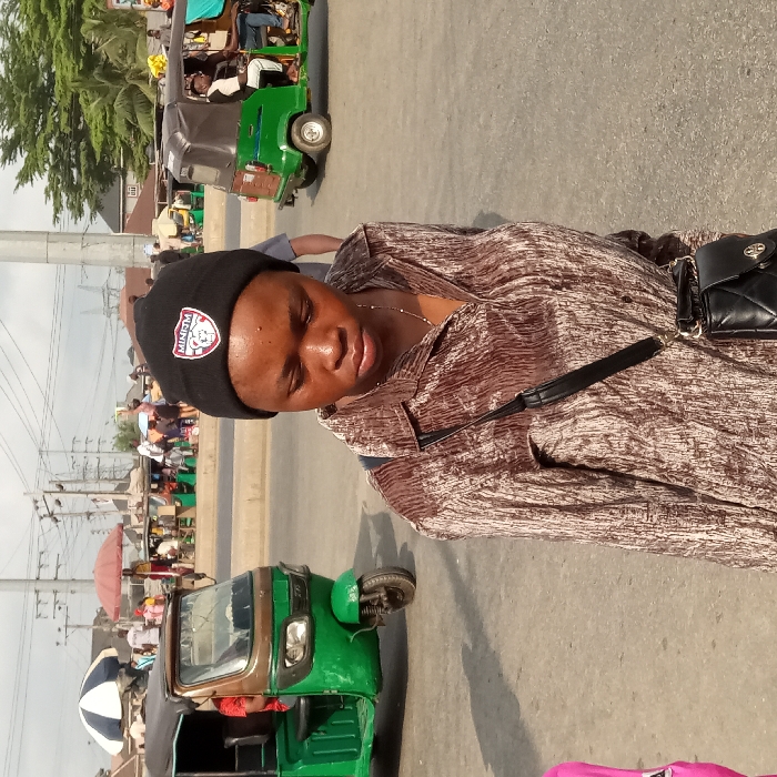 A person wearing a black beanie and brown patterned shirt stands on a street. Several green and black motorized tricycles, commonly known as "keke," are visible around her. A black handbag with a strap hangs on the person's shoulder. The background is a bustling street scene with people, power lines, and various buildings. The lighting indicates a sunny day.